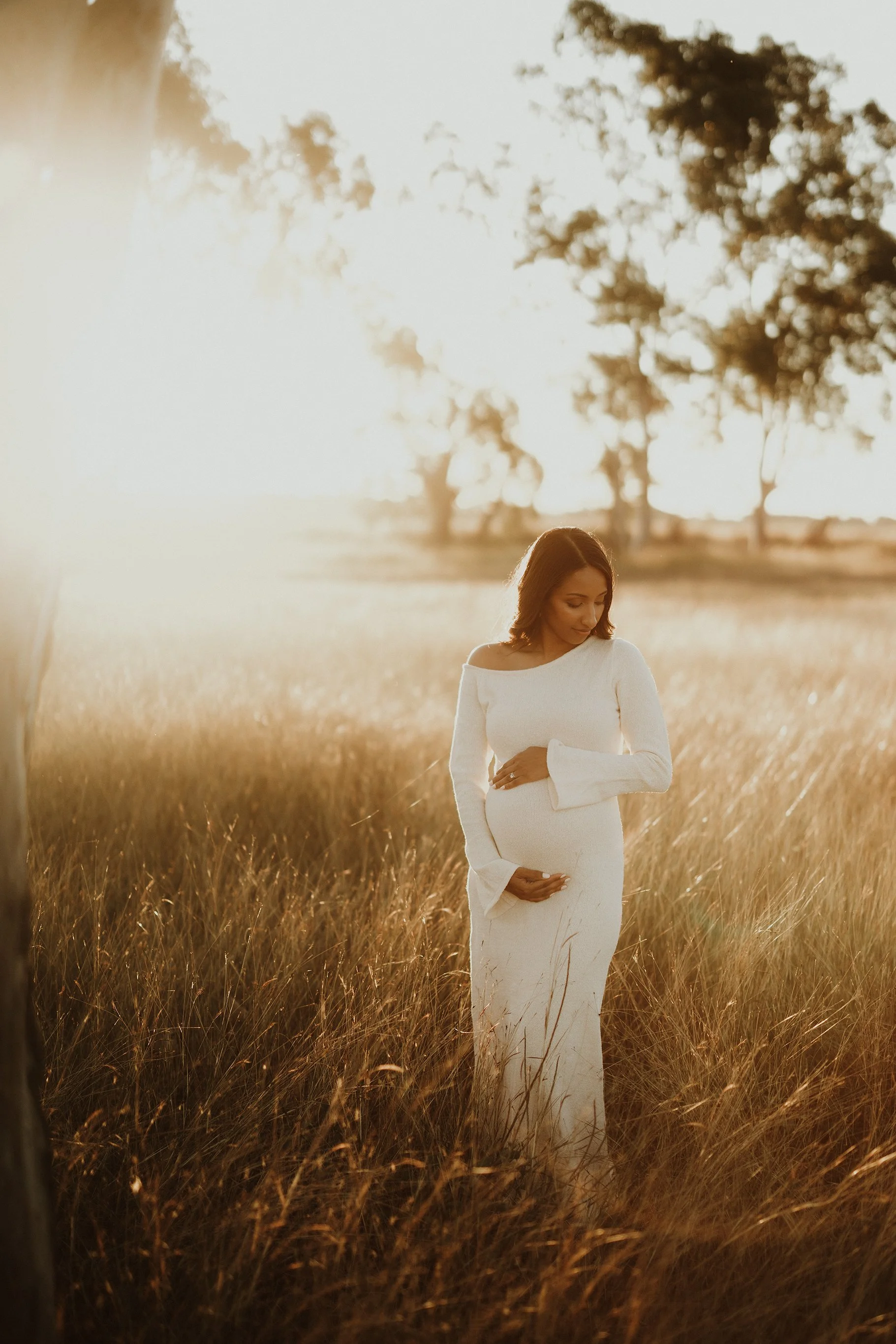 A pregnant woman wearing a white dress standing in a grassy field at sunset, gently holding her belly and looking down.