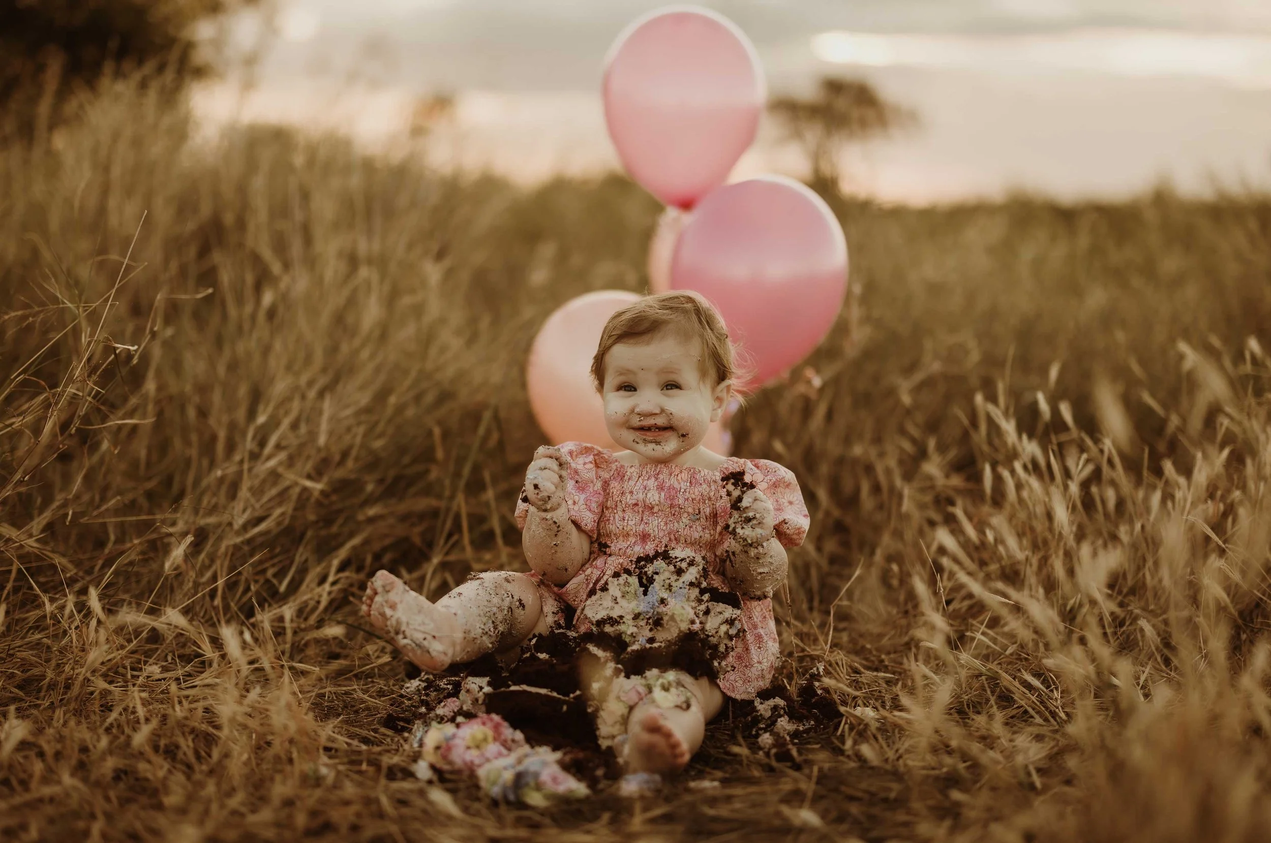 Baby sitting in a field of tall grass with pink balloons in the background, covered in cake and icing, celebrating a first birthday.