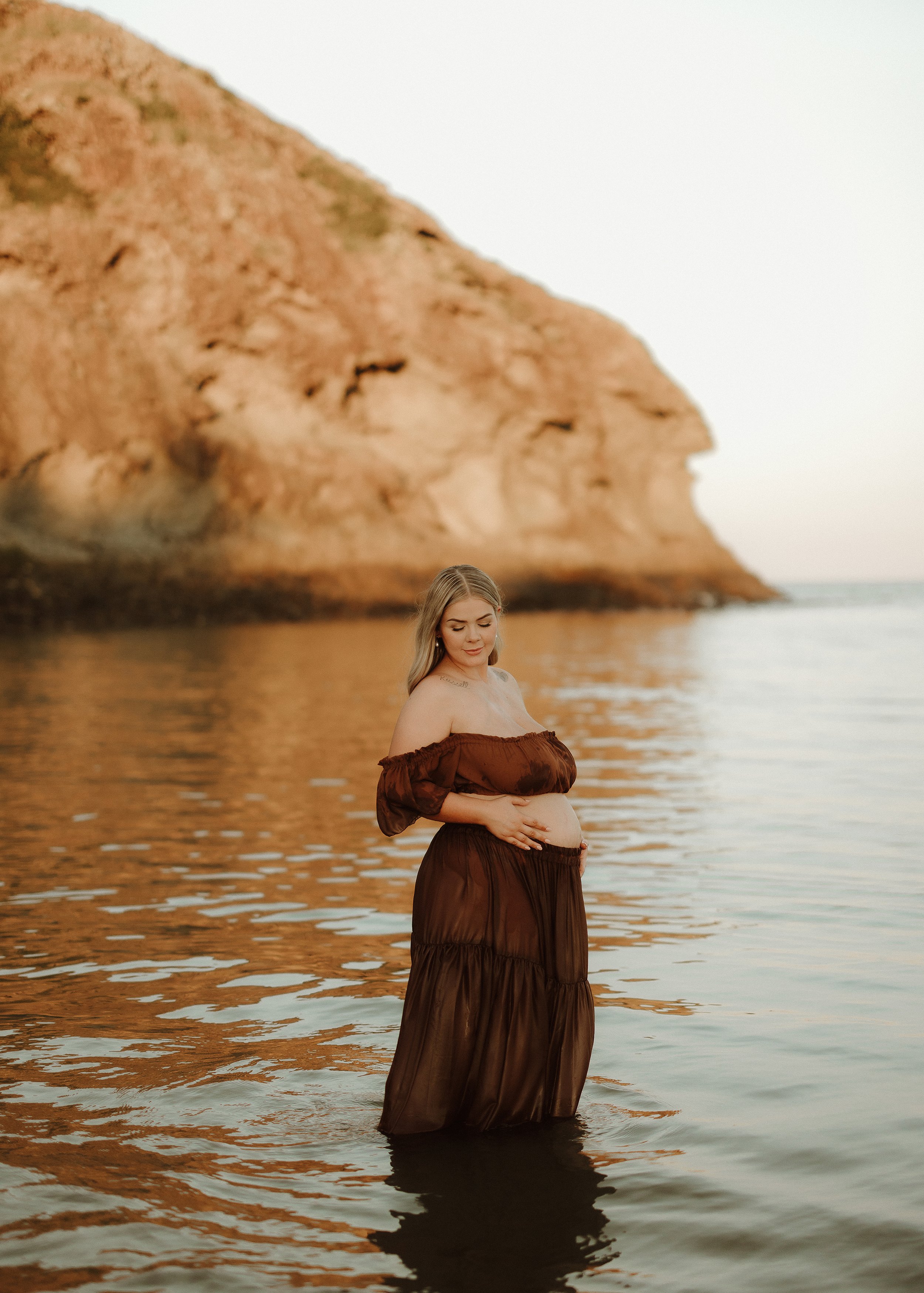 A pregnant woman standing in the water at the beach during sunset, wearing a flowing brown dress with a scenic rocky cliff in the background.