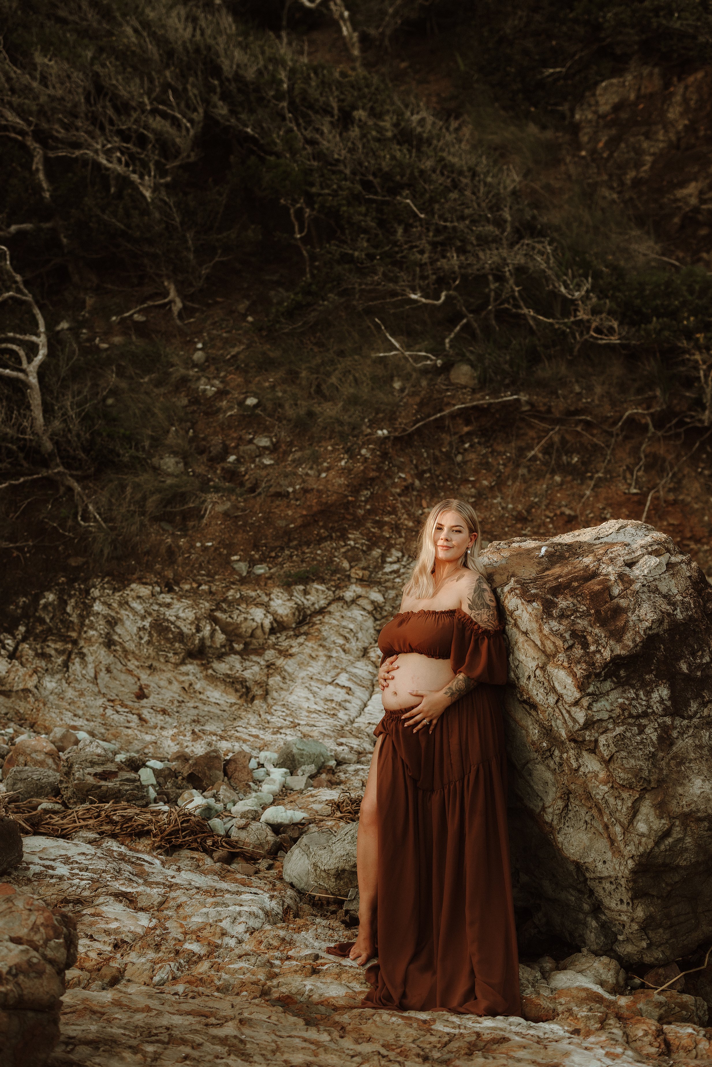 A pregnant woman in a brown off-the-shoulder dress standing on a rocky terrain against a large boulder and hillside with sparse trees.