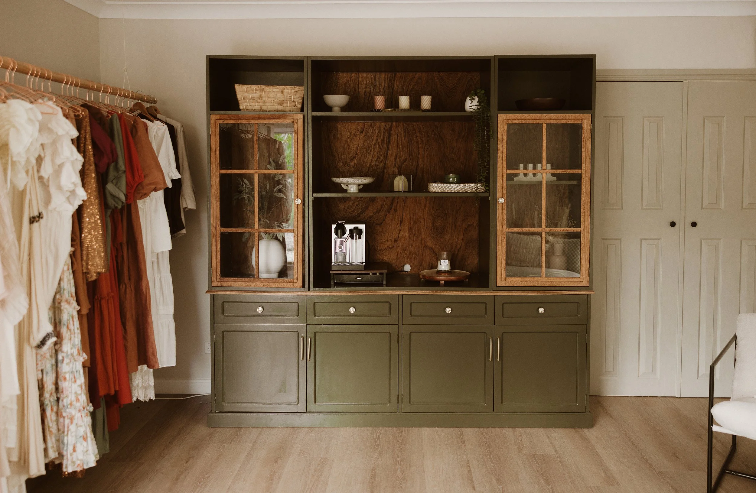 A vintage-style green and wood cabinet with glass-paned doors, empty shelves, and drawers in a room with hanging clothing on the left and a white door on the right.