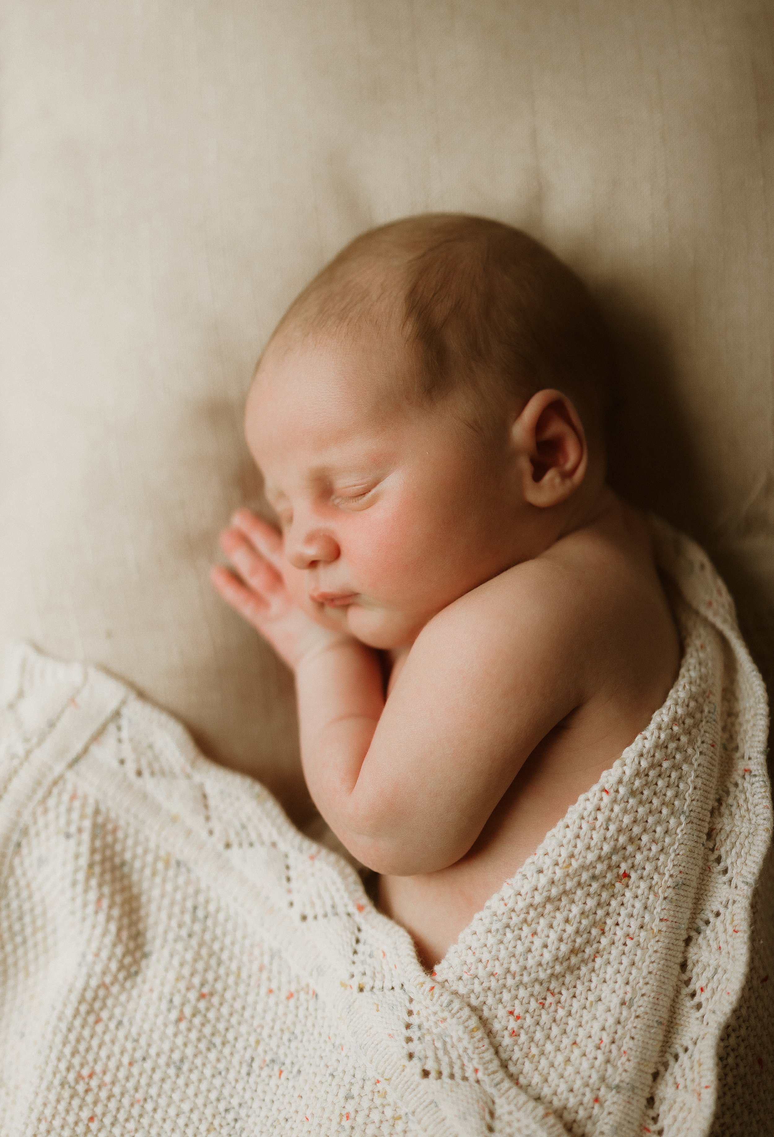 A sleeping newborn baby with closed eyes, lying on a beige surface, wrapped in a cream-colored knitted blanket.
