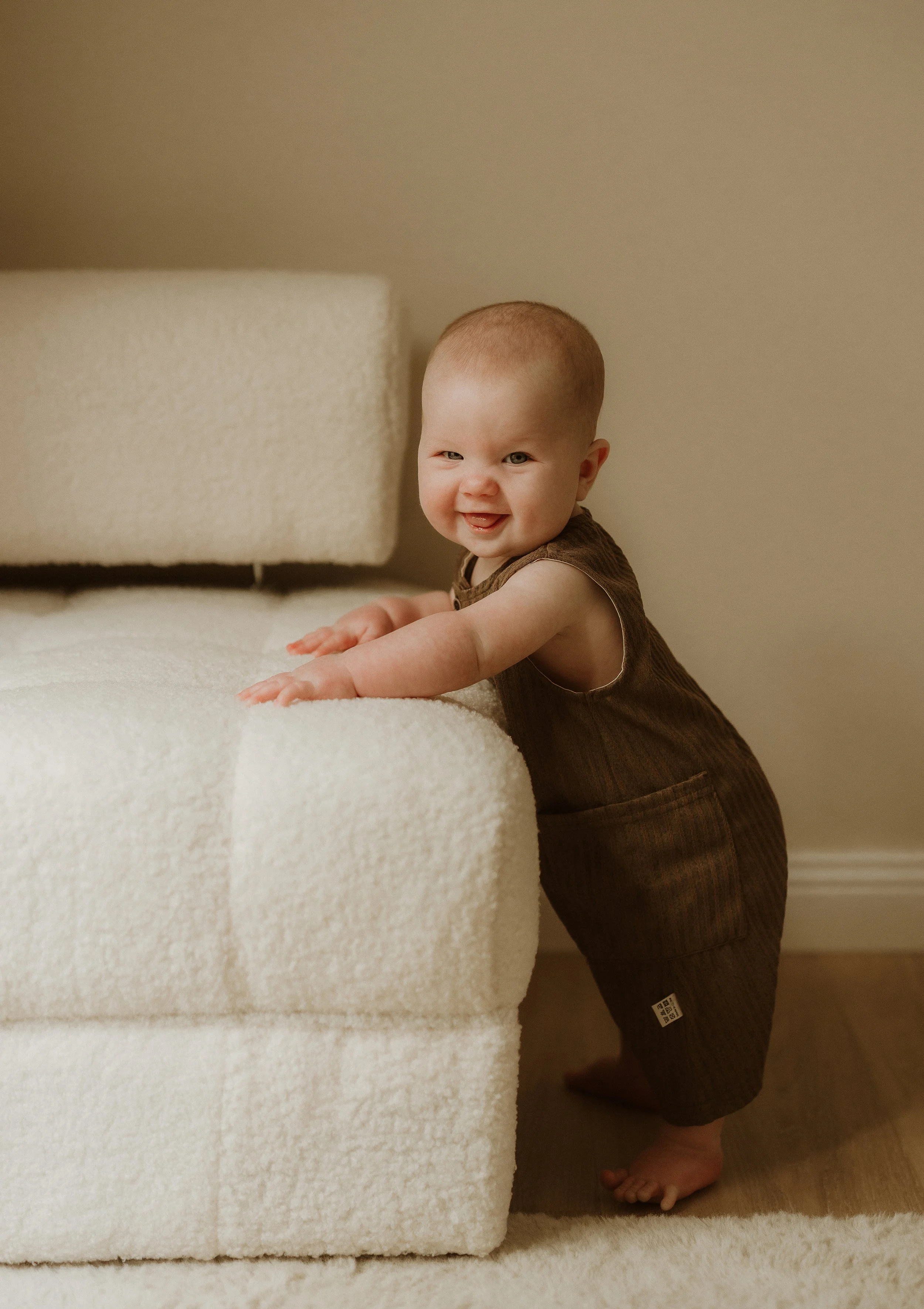 A smiling baby boy standing behind a cream-colored plush sofa, holding onto the edge and looking at the camera.