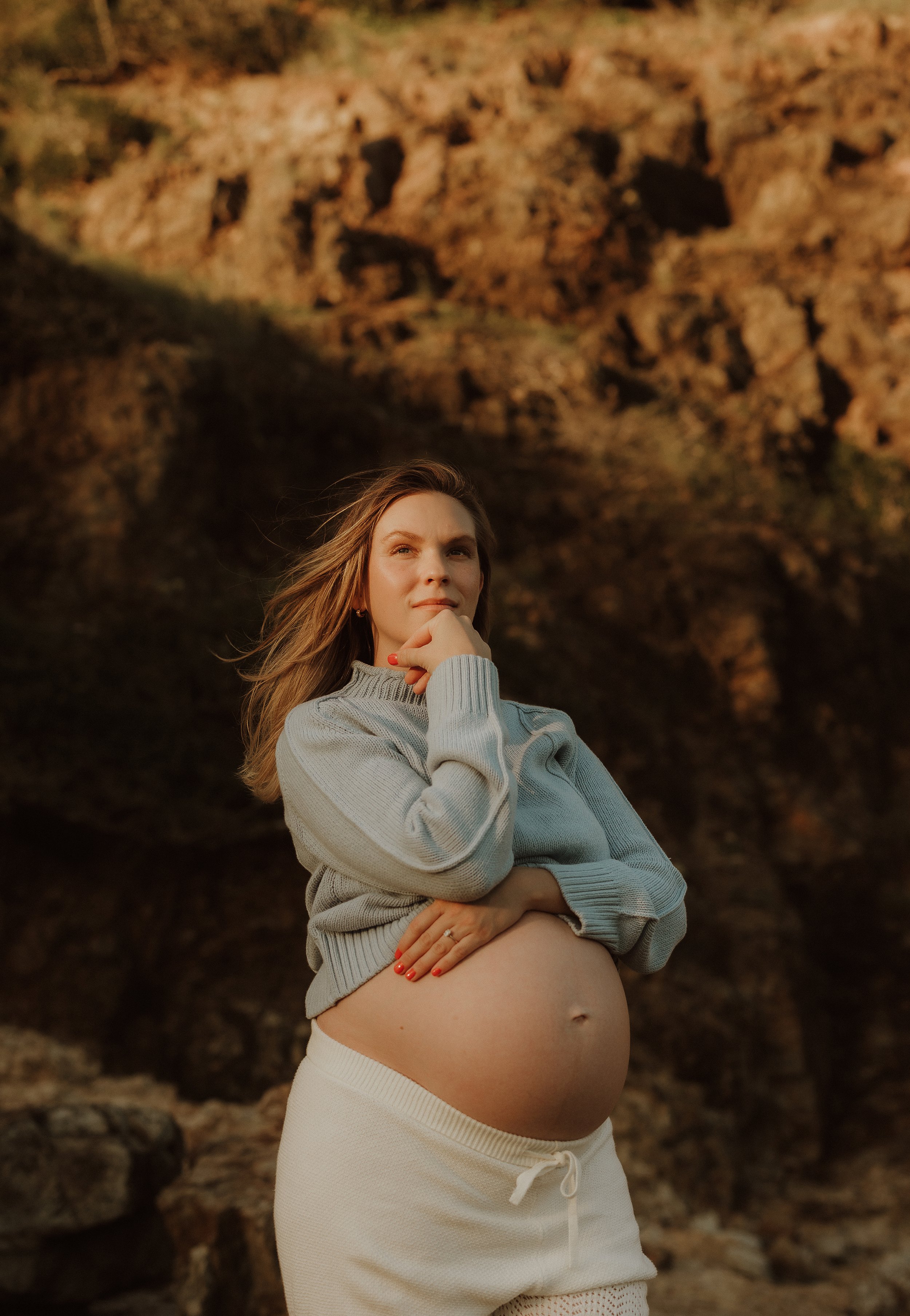 Pregnant woman in a gray sweater and cream sweatpants standing outdoors near rocks, with her hand on her chin and looking thoughtfully into the distance.