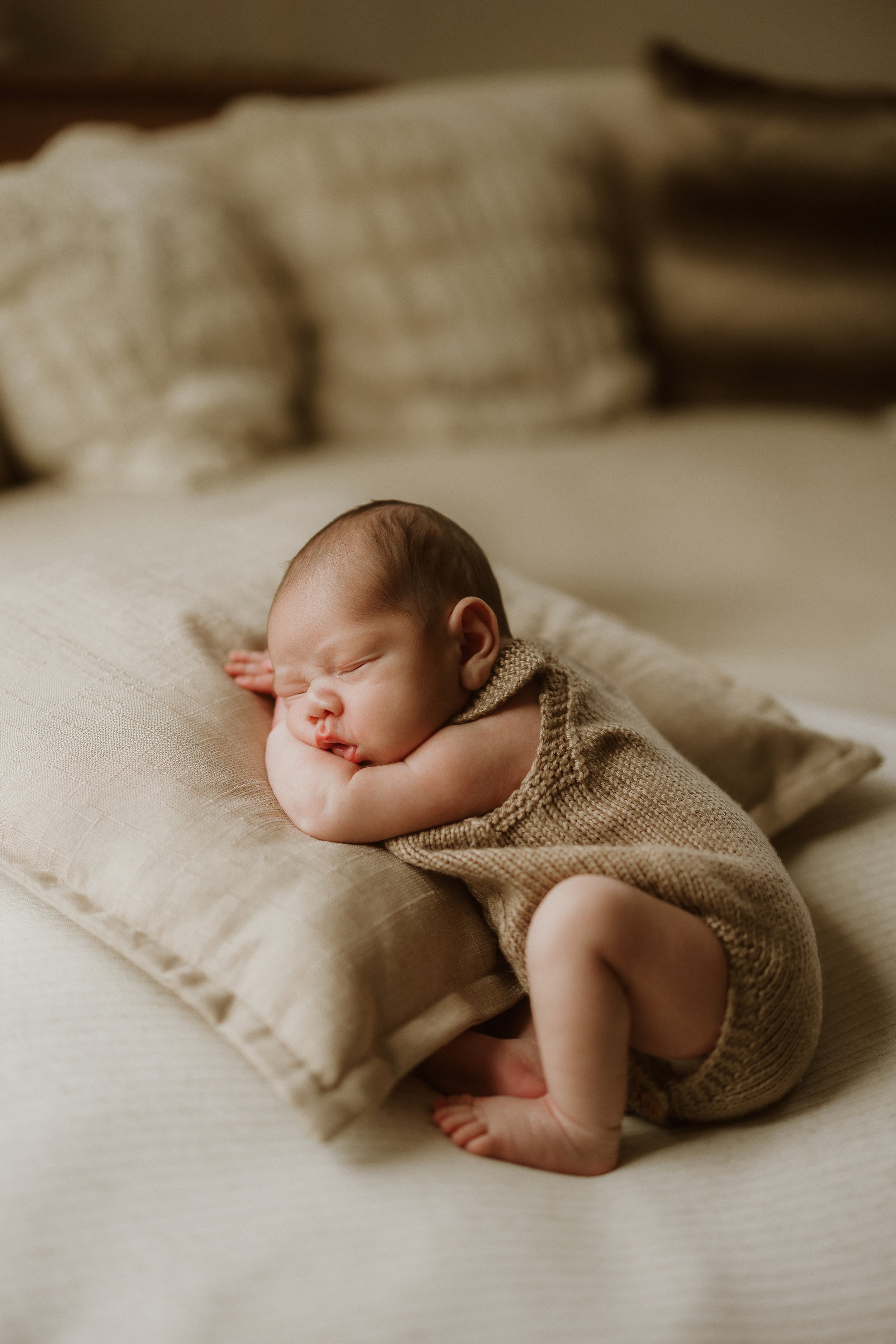 A sleeping newborn baby lying on a beige pillow on a bed, dressed in a knitted beige outfit.