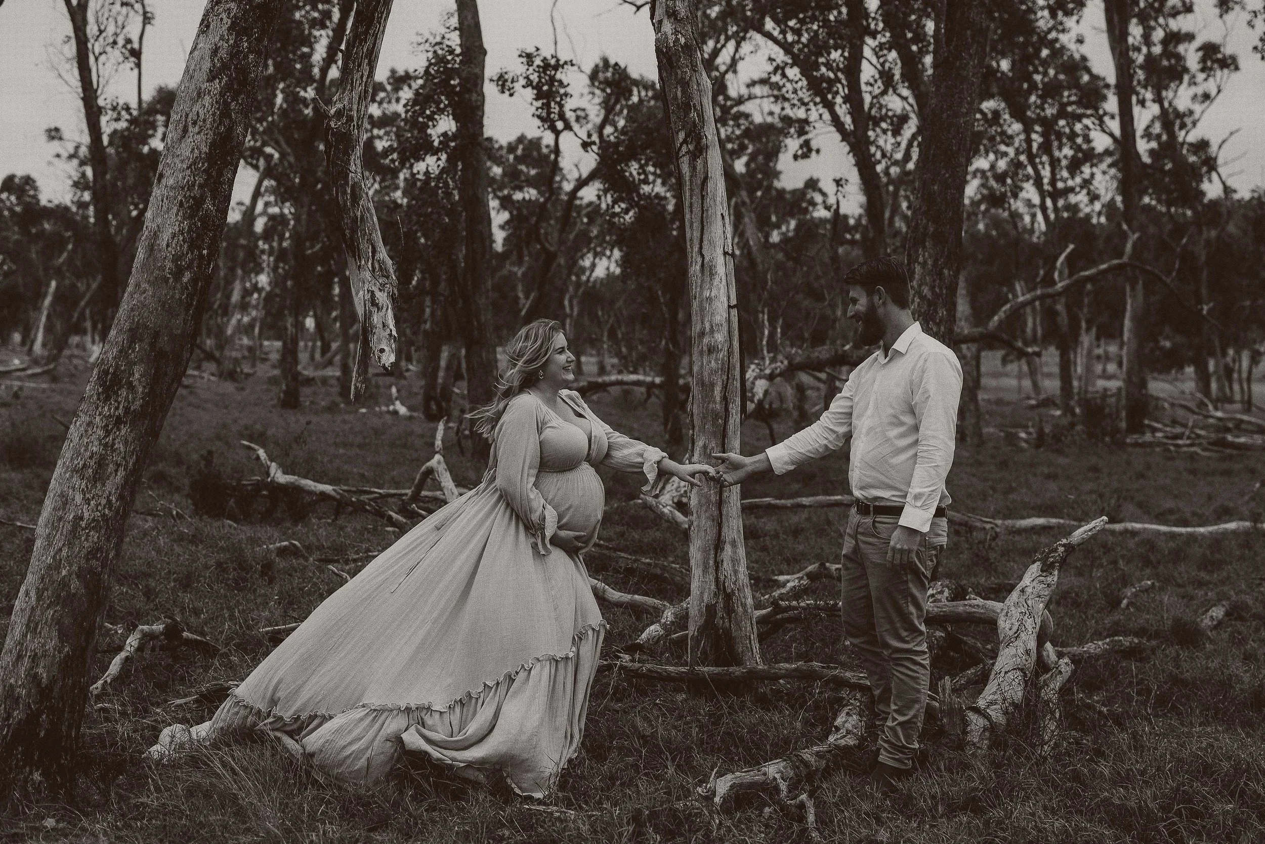 A couple holding hands in a forest, with a pregnant woman wearing a flowing dress and a man in a dress shirt and pants, black and white photo.
