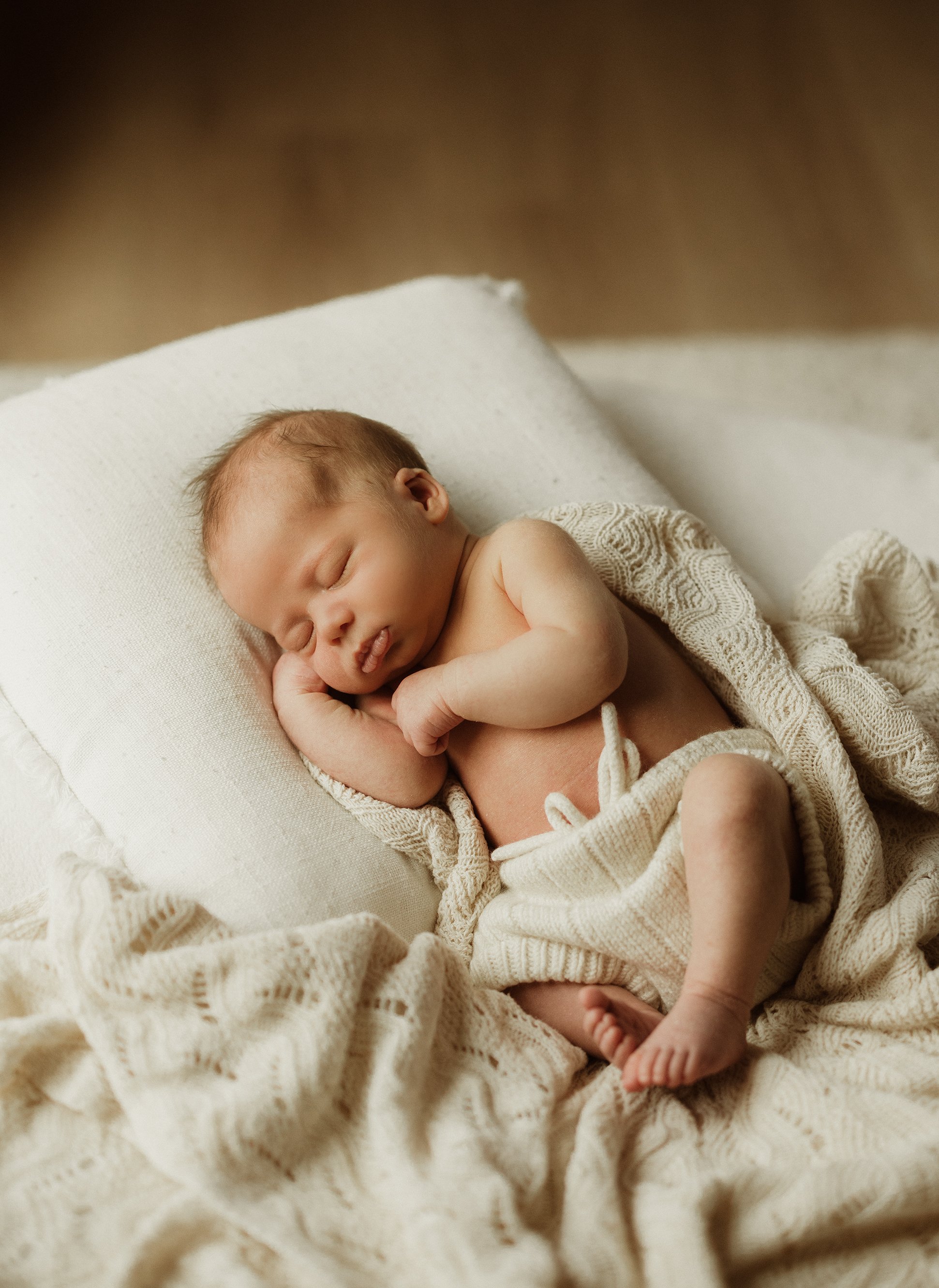 A sleeping newborn baby wrapped in a cream knitted blanket, lying on a white pillow and blanket.