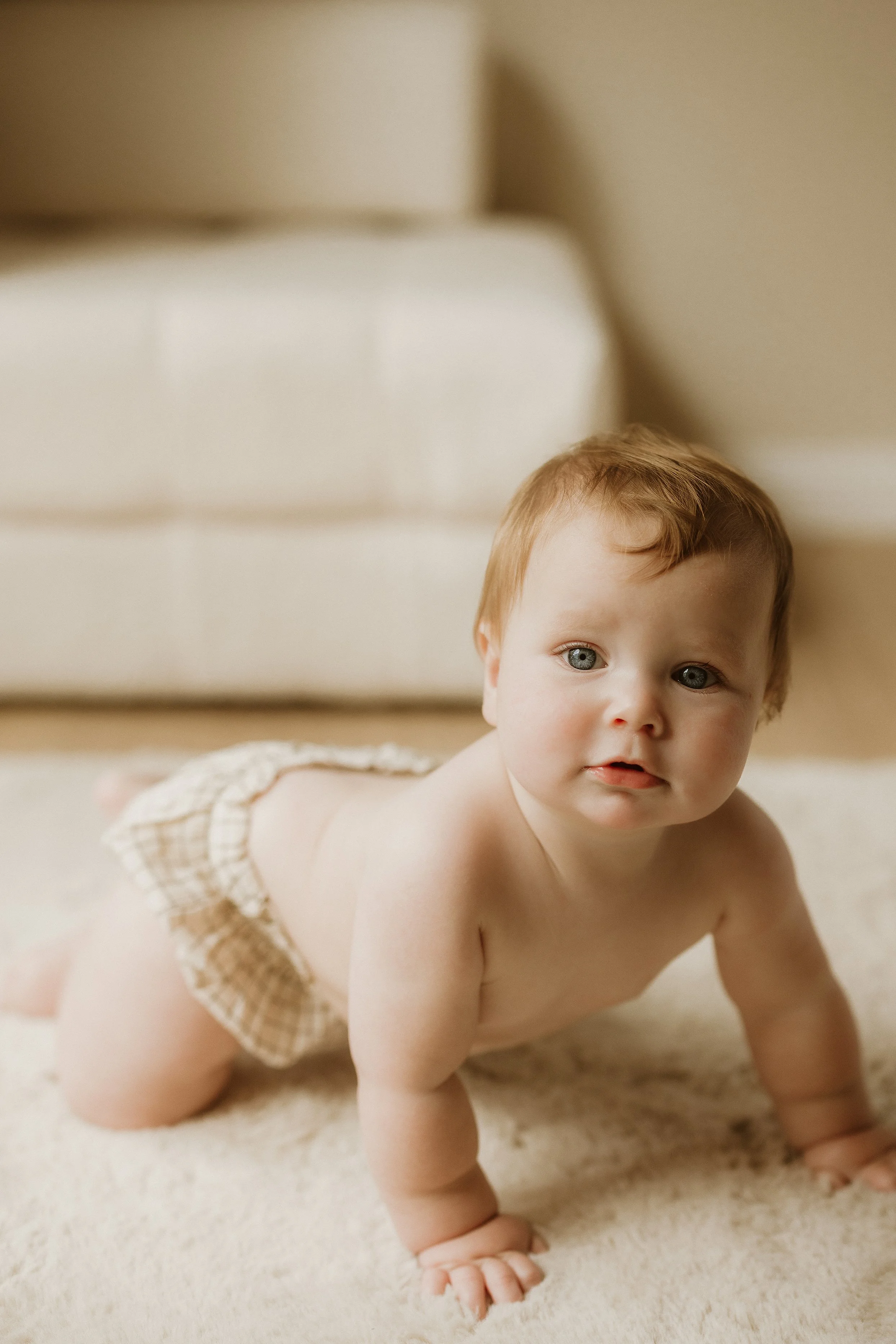 A baby with red hair crawling on a soft cream-colored carpet in a cozy room with beige furniture.