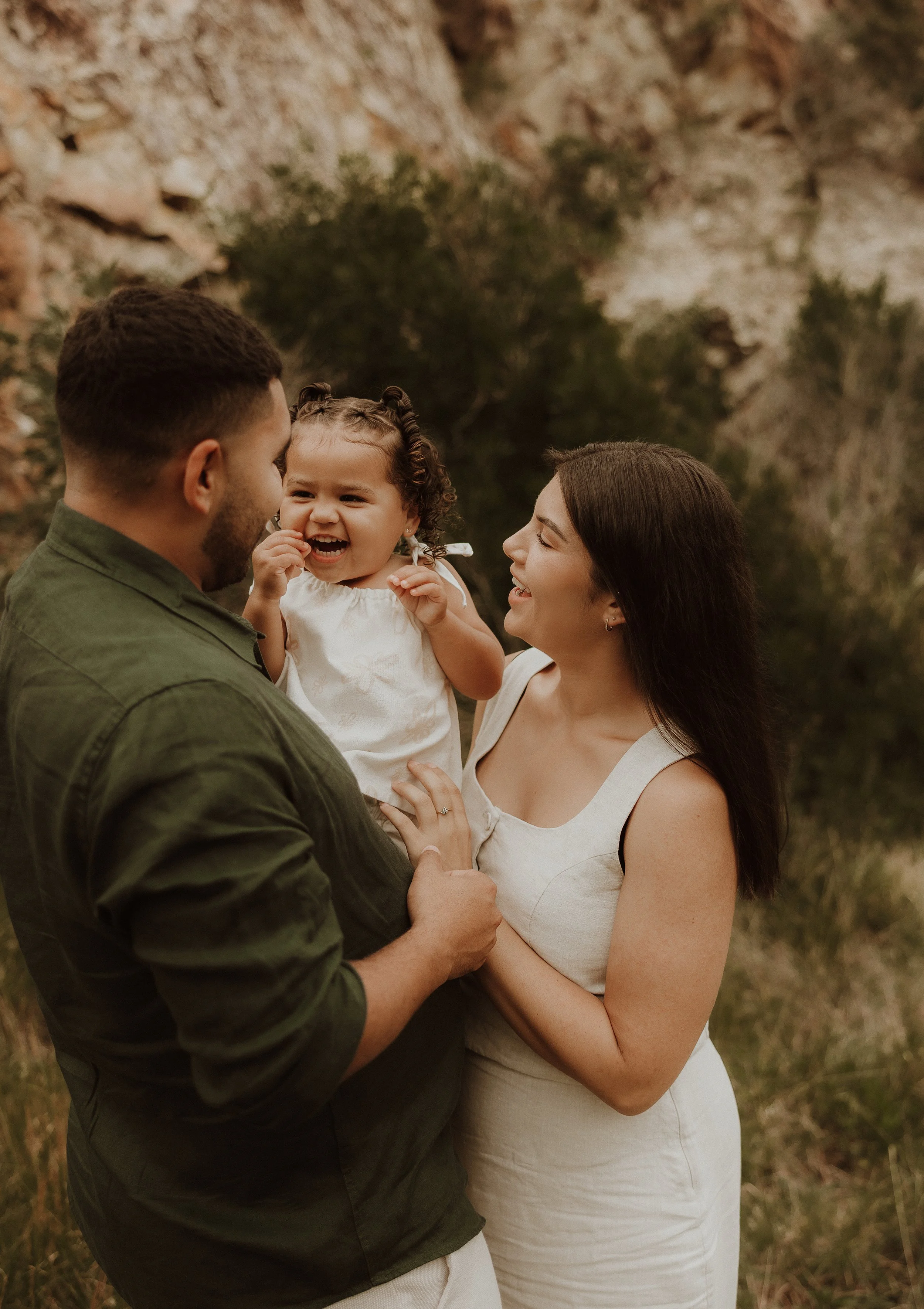 A happy family of three outdoors, with a father holding a young girl and a mother smiling at her, against a natural rocky background.