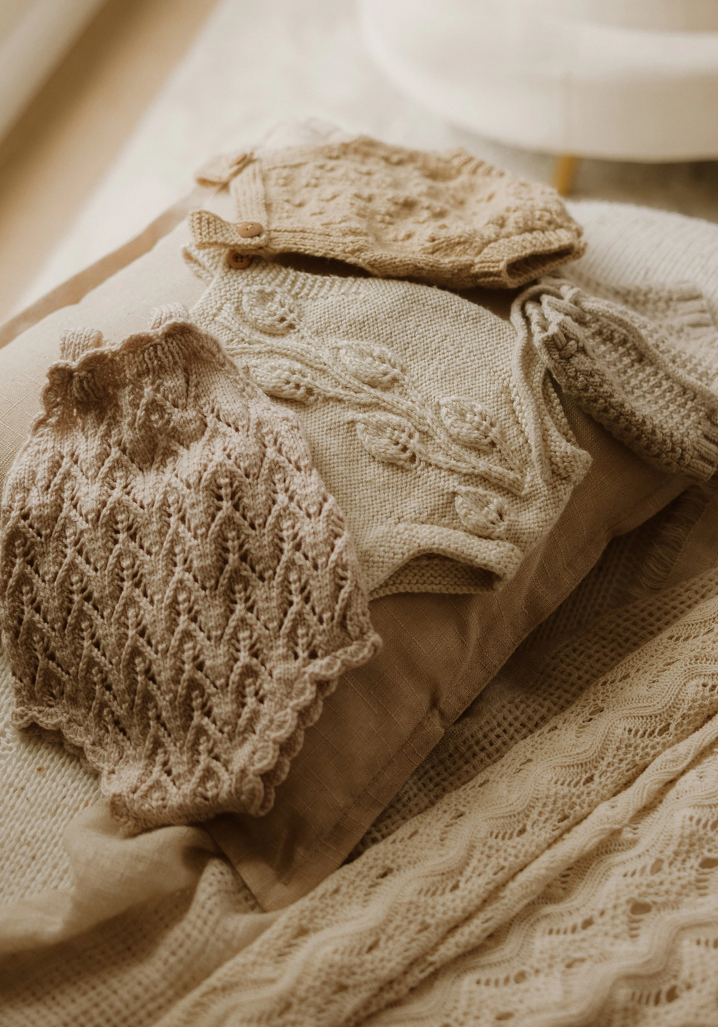 A collection of beige, cream, and light brown knitted and crocheted clothing items, including sweaters, socks, and other garments, arranged on a bed with beige and patterned linens.