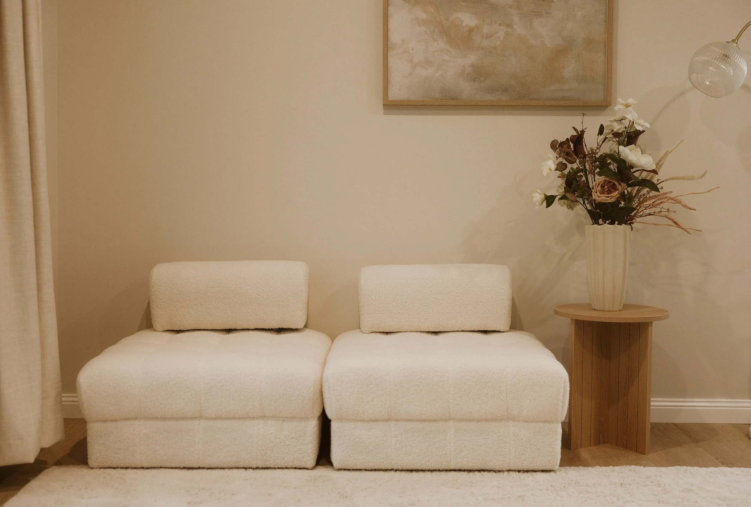 Living room with two white upholstered chairs, a wooden side table with a tall vase of flowers, artwork on the wall, and a lamp in a cozy, modern decor style.