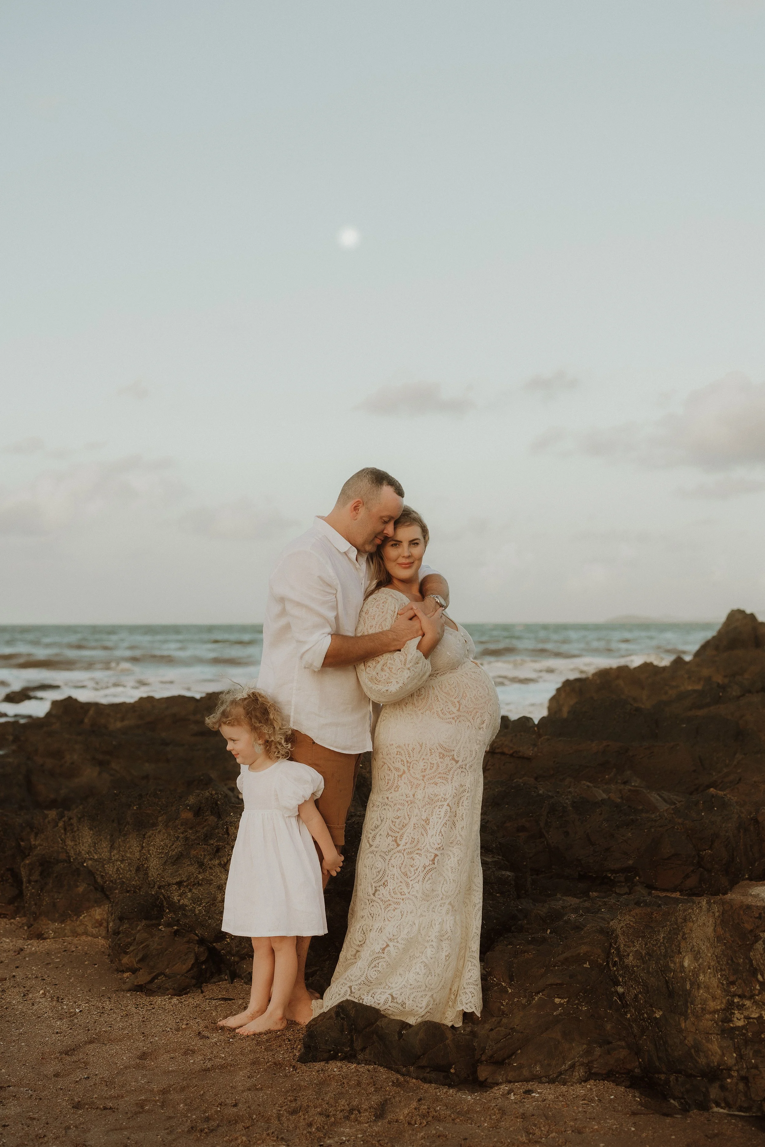 A pregnant woman and a man stand together on a rocky beach at sunset, with a young girl standing beside them, all dressed in white.
