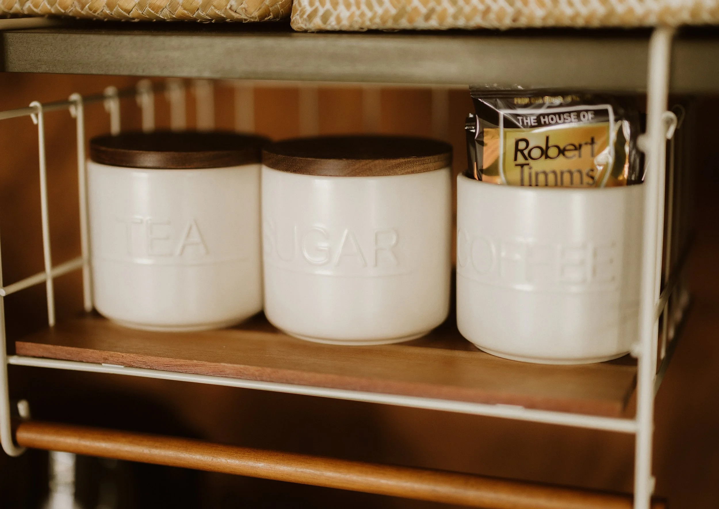 Cream-colored canisters labeled 'Tea', 'Sugar', and 'Coffee' on a wooden shelf inside a kitchen cabinet.