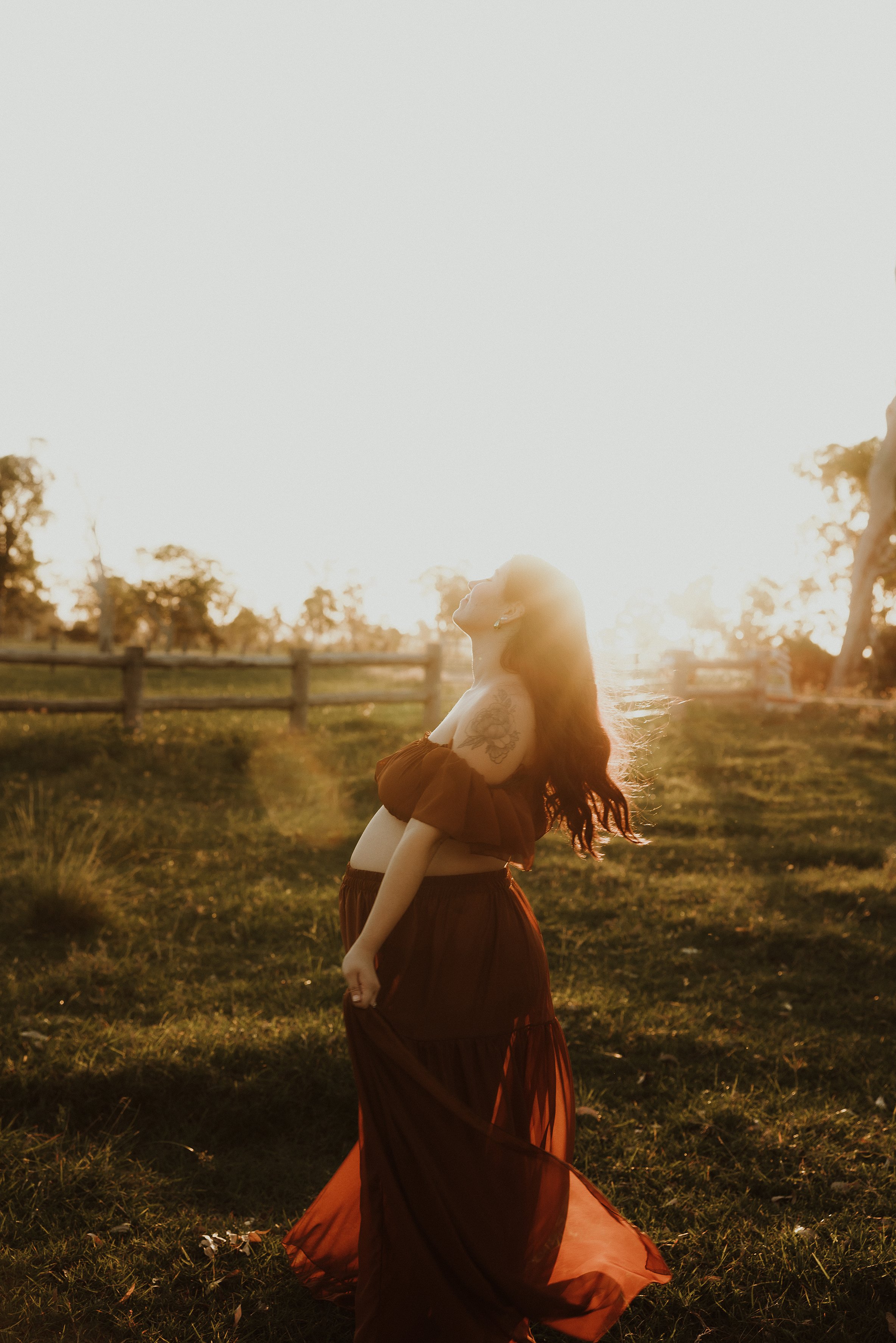 A pregnant woman in a flowing maroon dress standing outdoors at sunset, with her head tilted back and hair flowing, near a wooden fence and trees in the background.