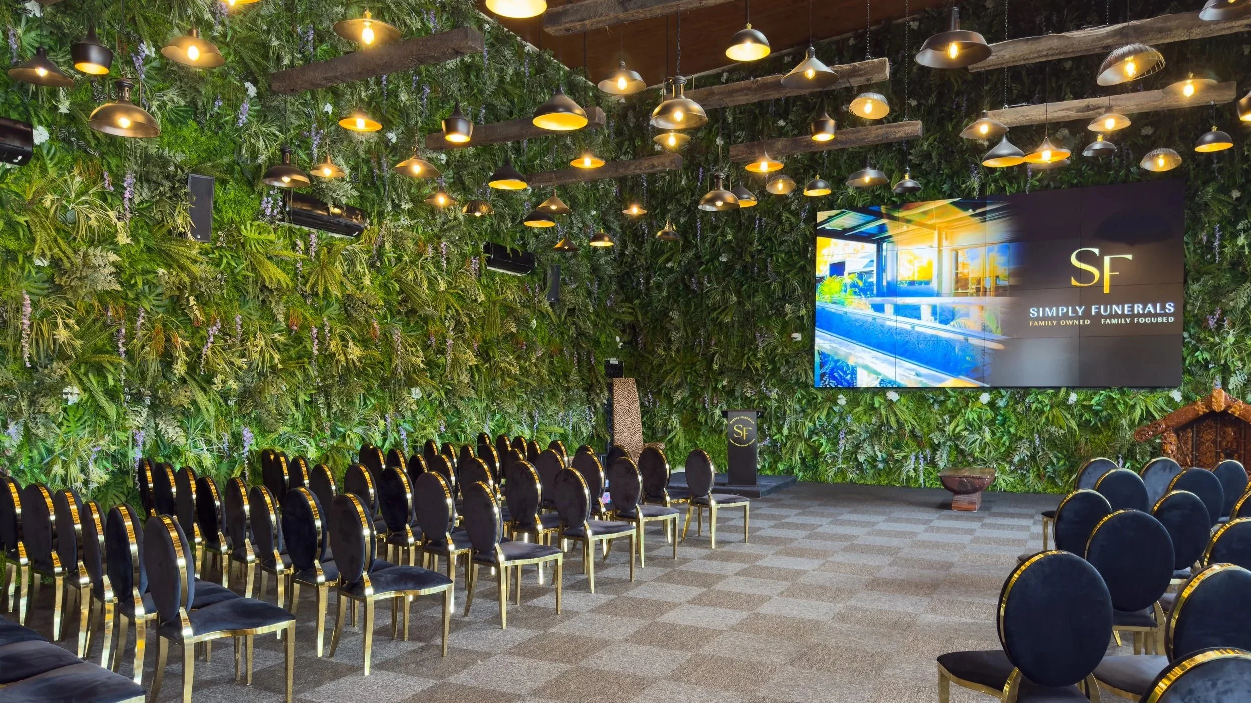 Indoor funeral home event space with black chairs arranged in rows, green living wall backdrop with plants, wooden ceiling with hanging lights, a large digital sign displaying 'SF Simply Funerals Family Owned | Family Focused', and a small decorative