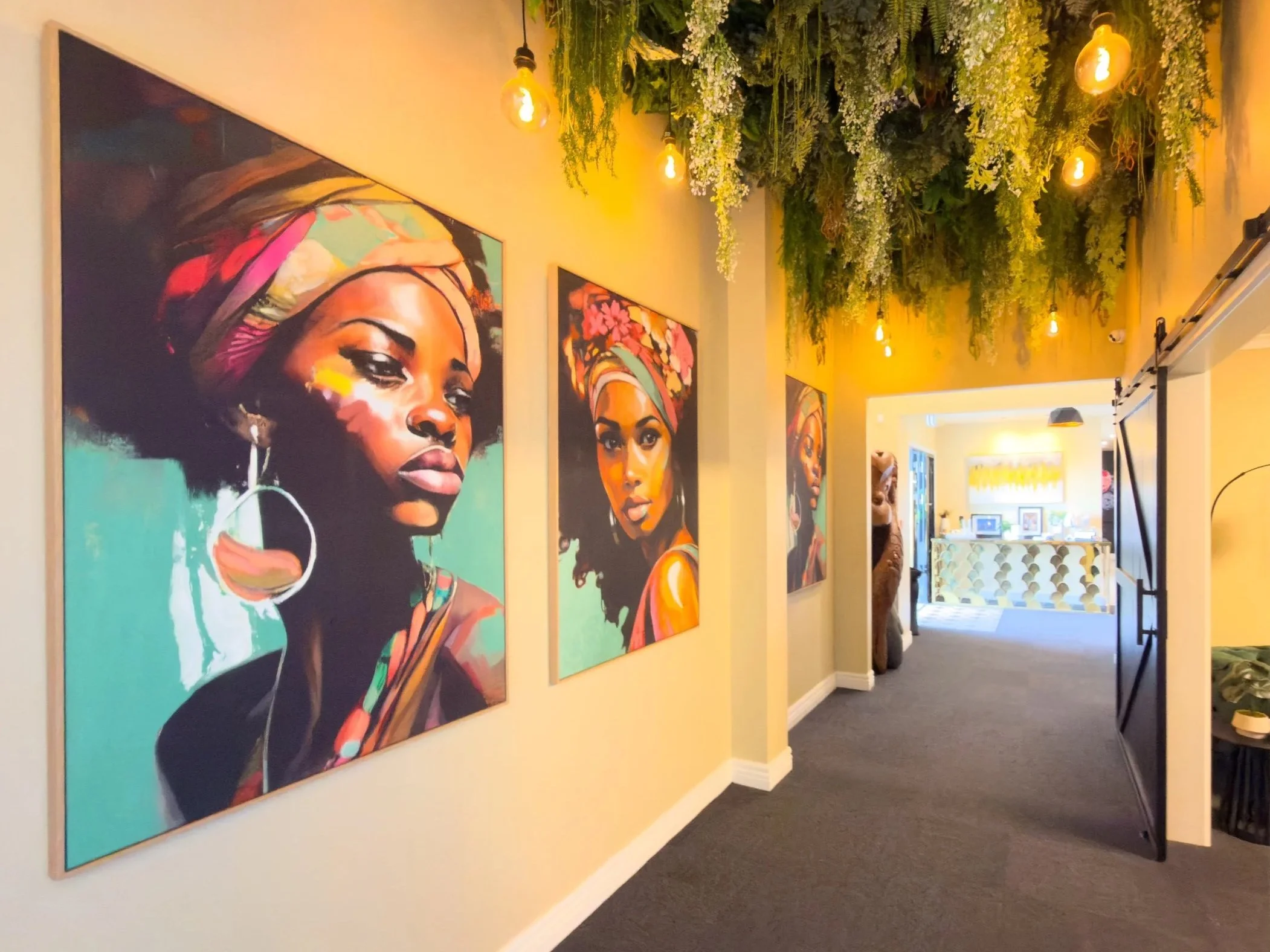 Interior hallway with hanging greenery ceiling decoration and Edison bulb lighting, featuring three large portrait paintings of Black women on the left wall, and a reception desk at the end of the hallway.