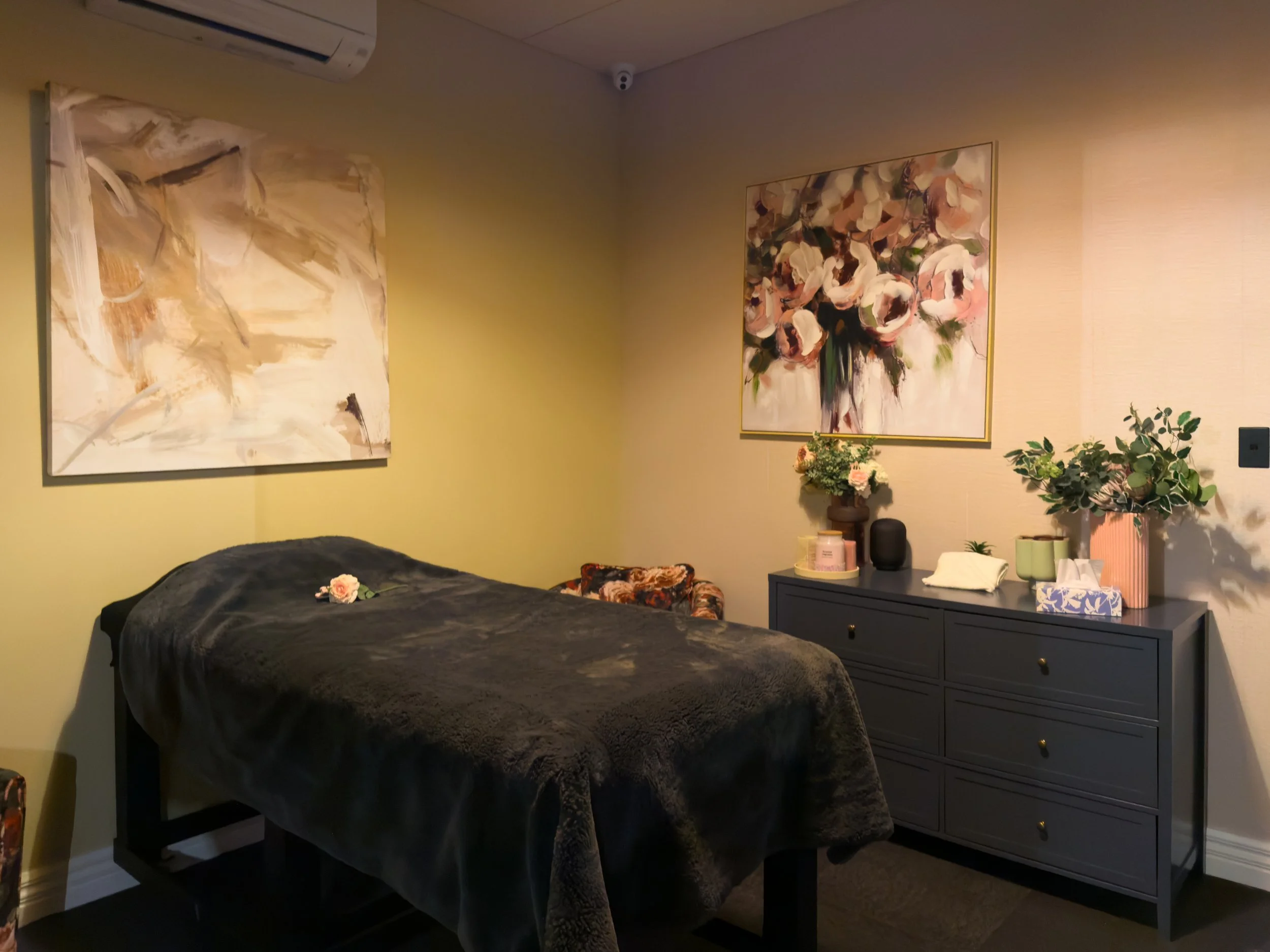 A cozy visitor room with a massage table, dark shoes, a blue couch, abstract wall art, a sideboard with flowers and candles, and warm lighting.