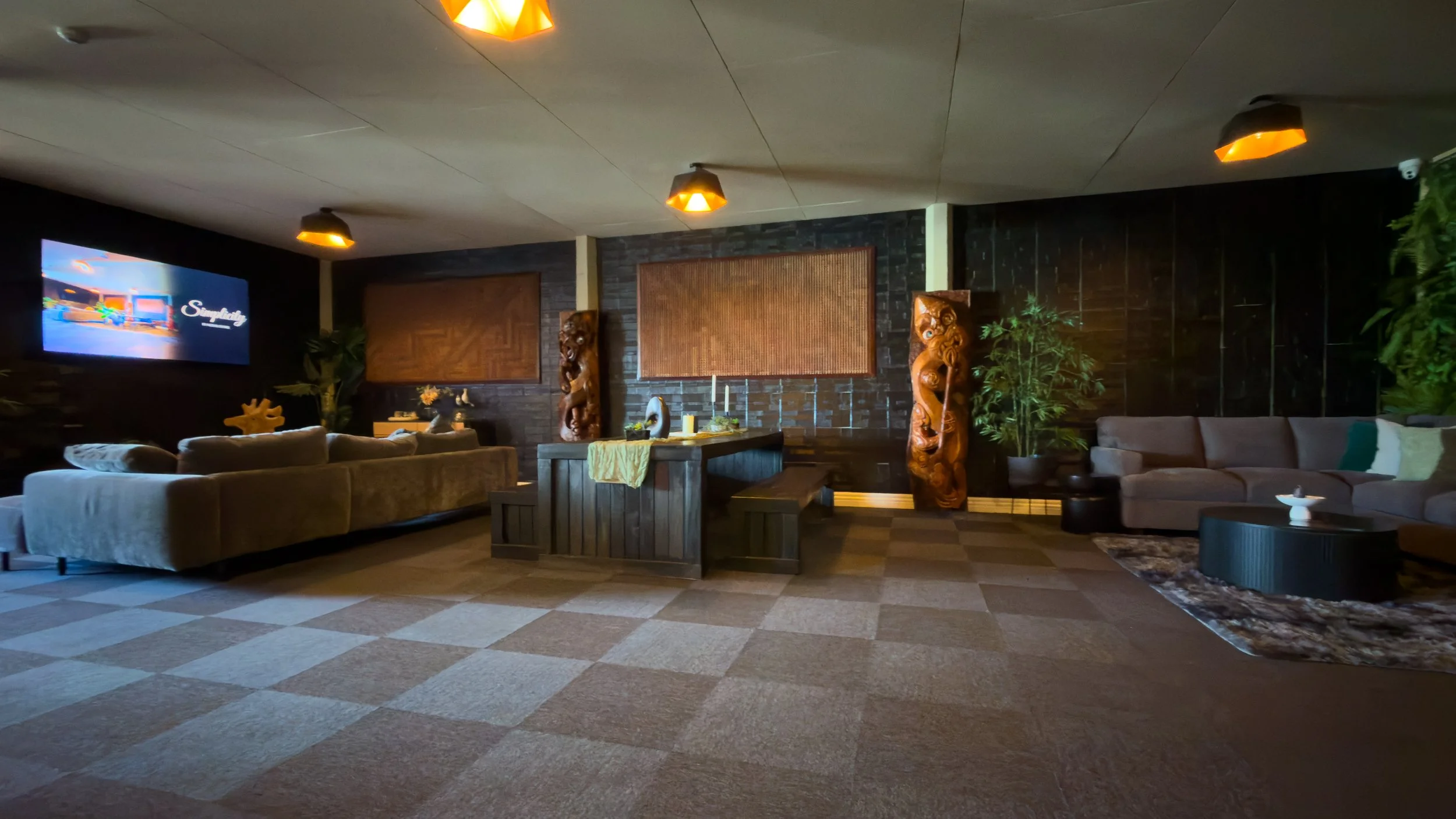 Our spacious vistor lounge with modern decor, including beige and tan sofas, wooden furniture, the wall decorated with dark bricks, tropical plants, and wooden sculptures, with ceiling lights and candles for illumination.