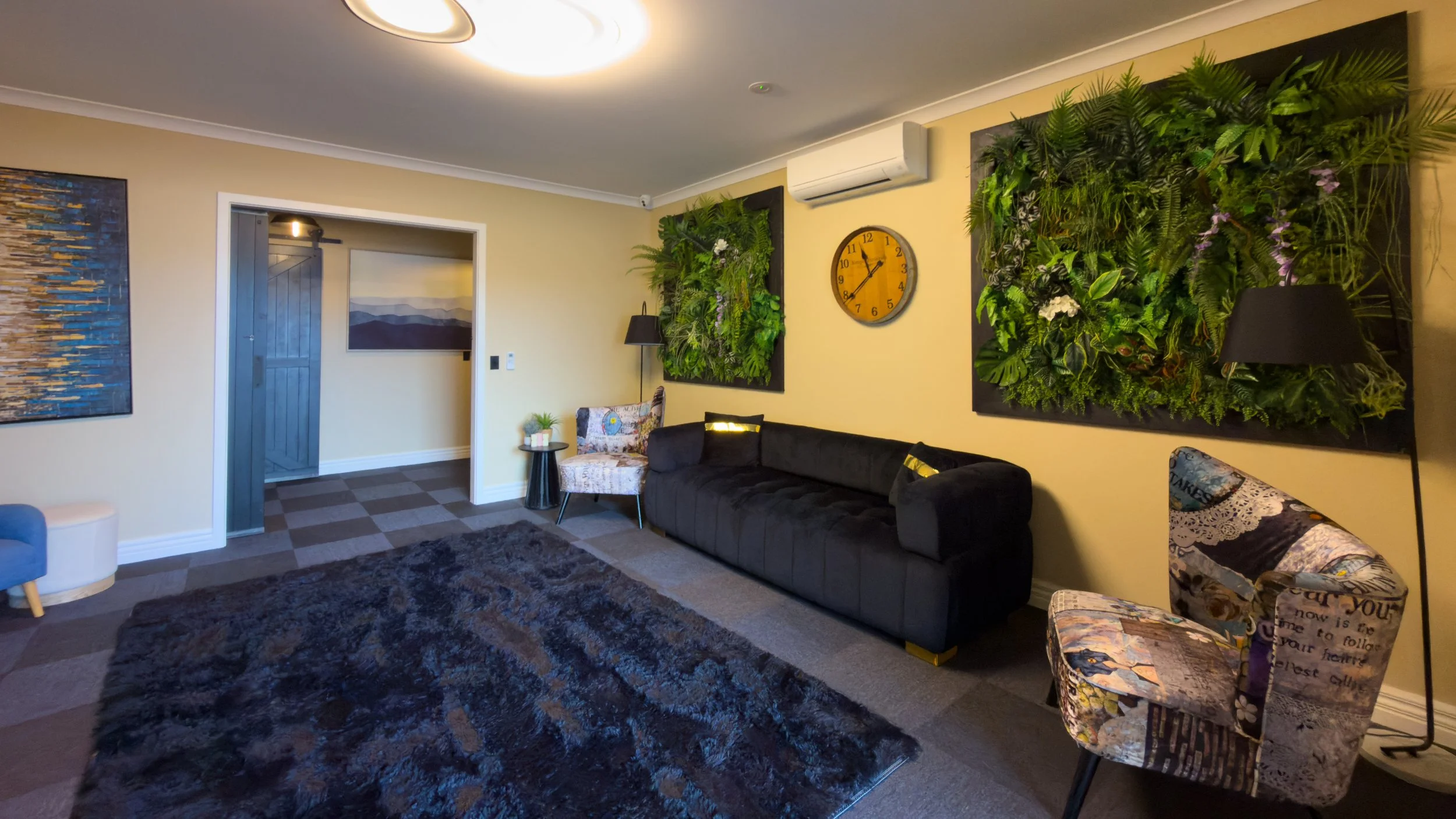 A spacious, modern  room with neutral-colored walls, dark wood barn doors, a large black rug, and various seating options including a gray armchair and a patterned sofa. There are decorative paintings on the walls, a black floor lamp, and some plants