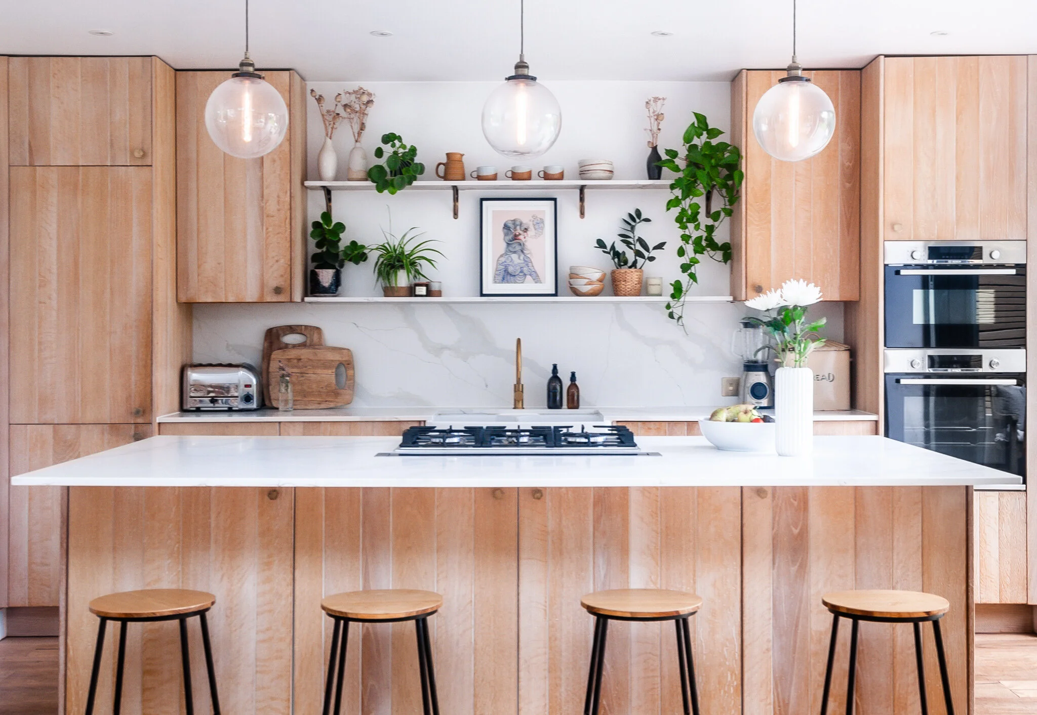 Light oak kitchen with island, open shelving and pendant lighting designed by Suellen Farias Interiors.