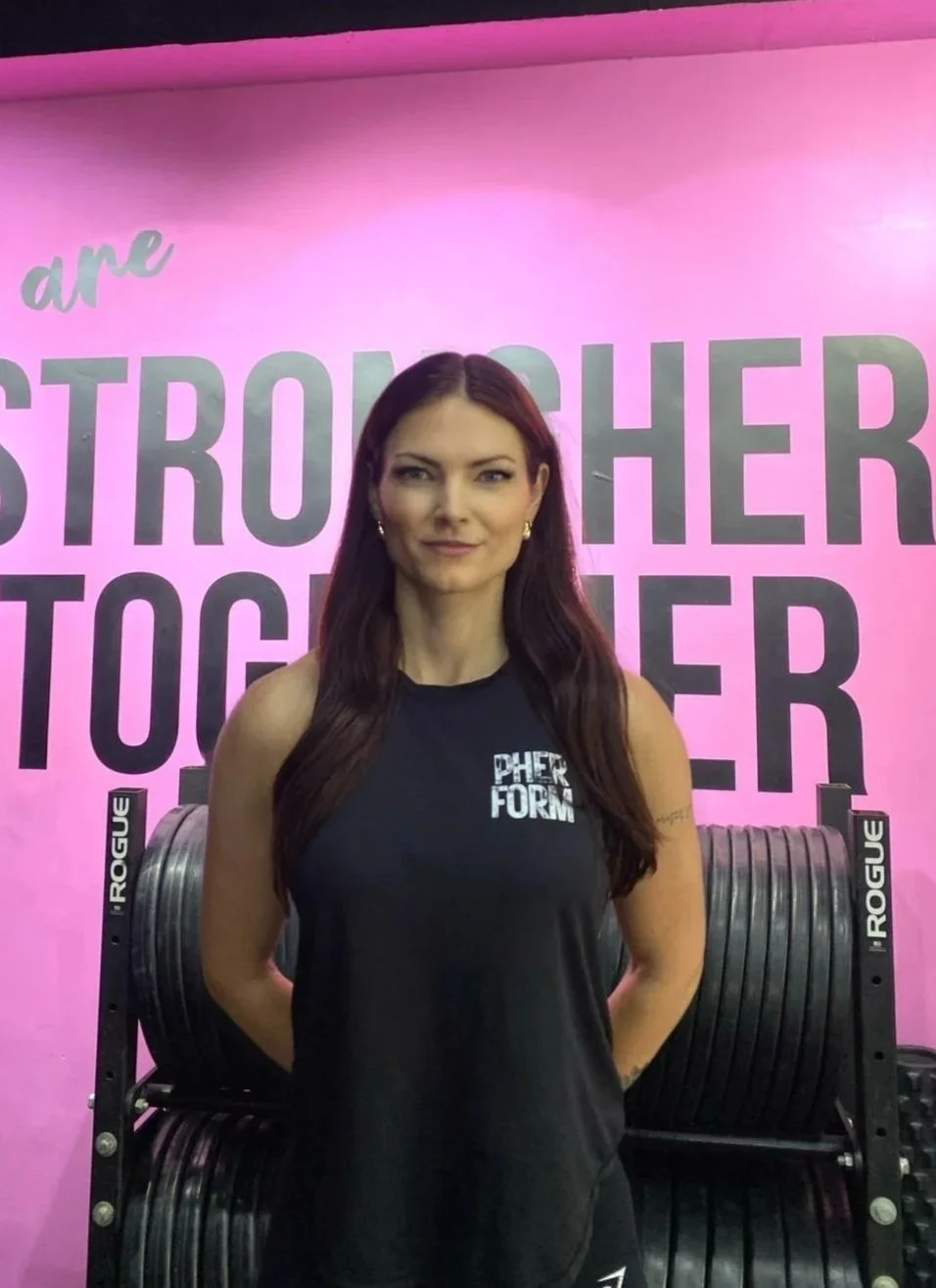 A woman with long brown hair smiling and crossing her arms, standing in front of a pink and purple background. She is wearing a black sleeveless top with the word 'pherform' in pink and black workout leggings, with fitness weights in the background.