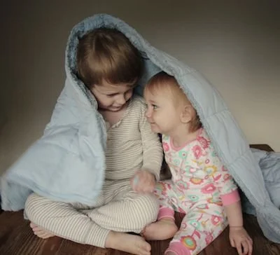 Two young children, a boy and a girl, sitting on the floor under a blanket draped over them, smiling and looking at each other.