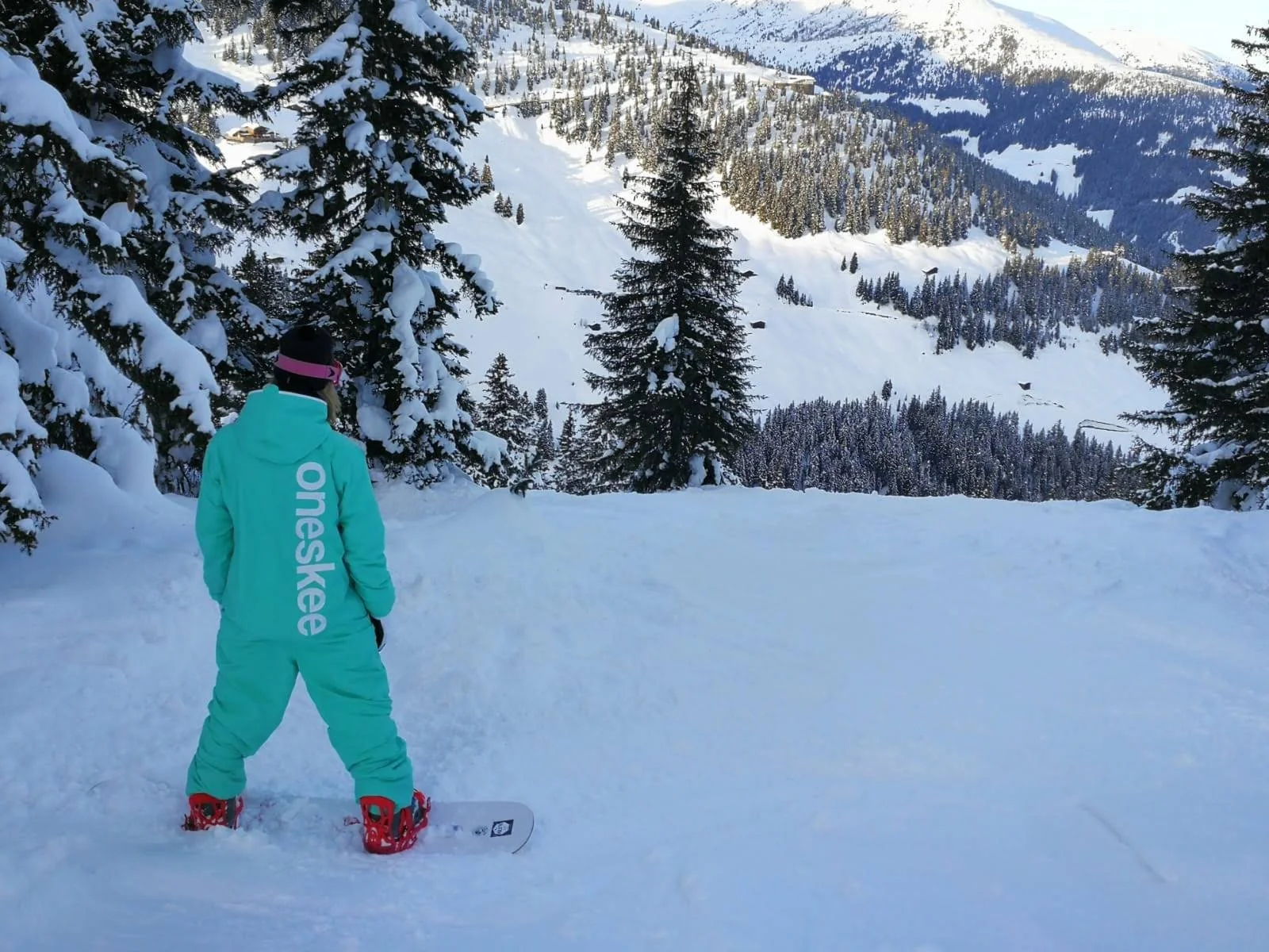 Eine Person in einer hellblauen Skihose und Jacke mit der Aufschrift 'Oneway' steht auf verschneitem Boden und blickt auf eine verschneite Winterlandschaft mit Tannenbäumen und Bergen im Hintergrund.