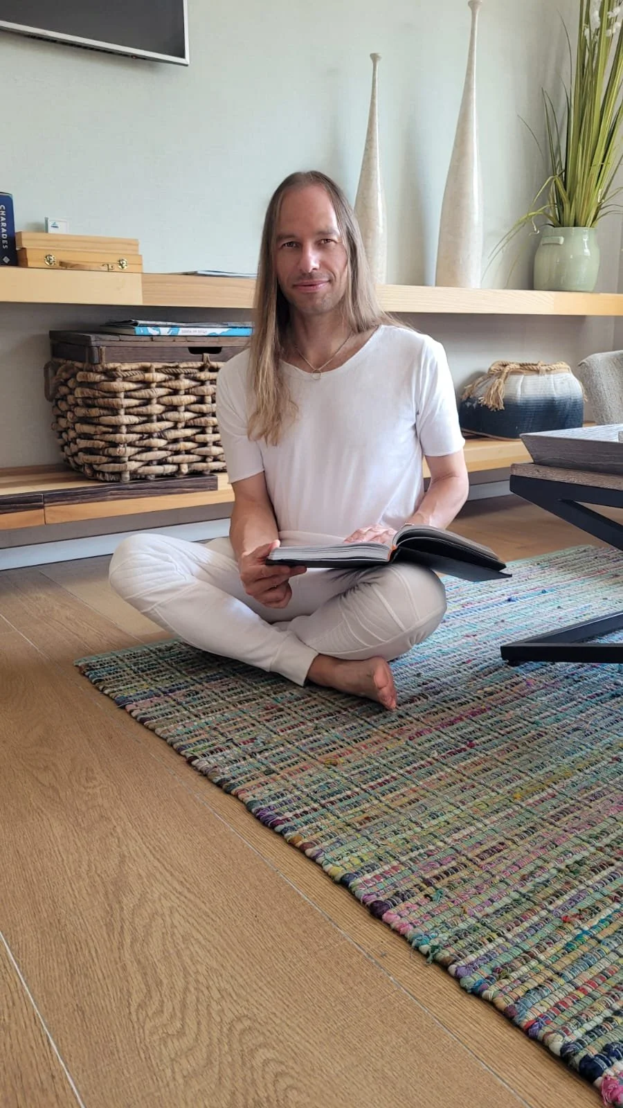 Ein Mann sitzt barfuß auf einem bunten Teppich auf dem Boden, liest ein Buch in einer modernen, stilvollen Wohnung.