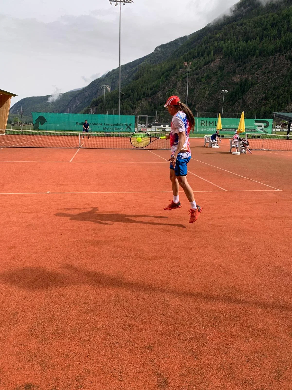 Sascha Geipel spielt Tennis auf einem roten Tennisplatz. Er trägt eine rote Kappe, ein buntes T-Shirt und blaue Shorts. Im Hintergrund sind Bänke mit Sonnen- oder Regenschirmen sowie andere Spieler. Es gibt Berge und Wolken im Hintergrund.