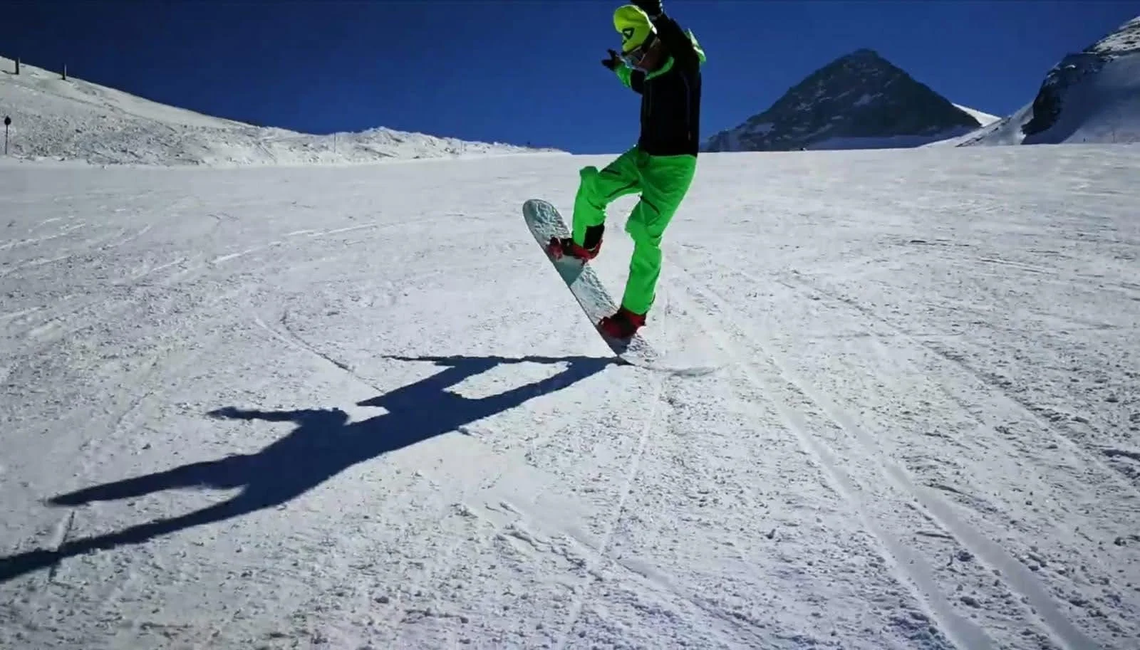 Skifahrer in grüner Kleidung springt auf schneebedecktem Hang bei klarem blauen Himmel, Berge im Hintergrund.