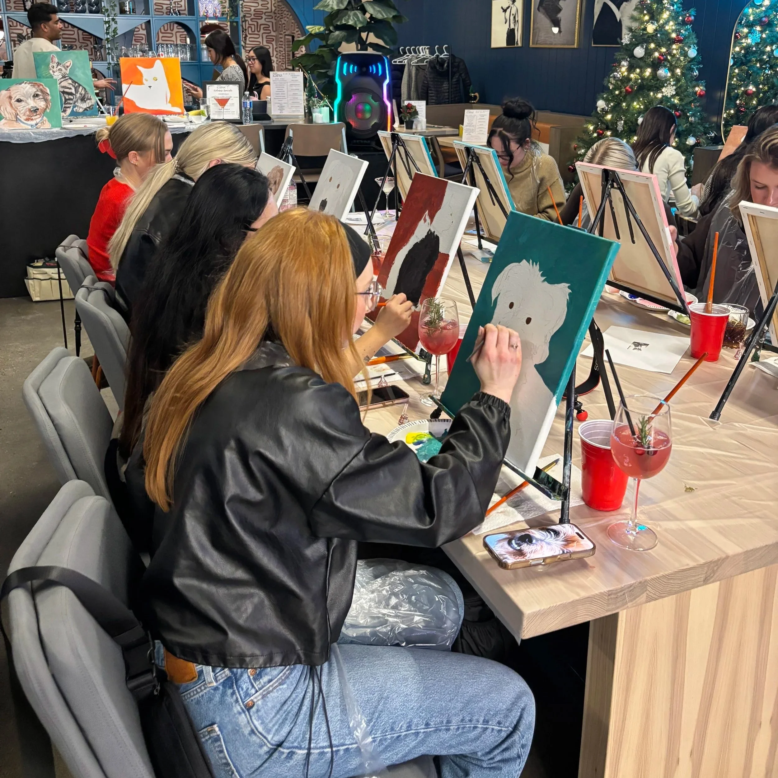 People participating in a group painting class at a restaurant or studio, with Christmas trees and art supplies on the tables.