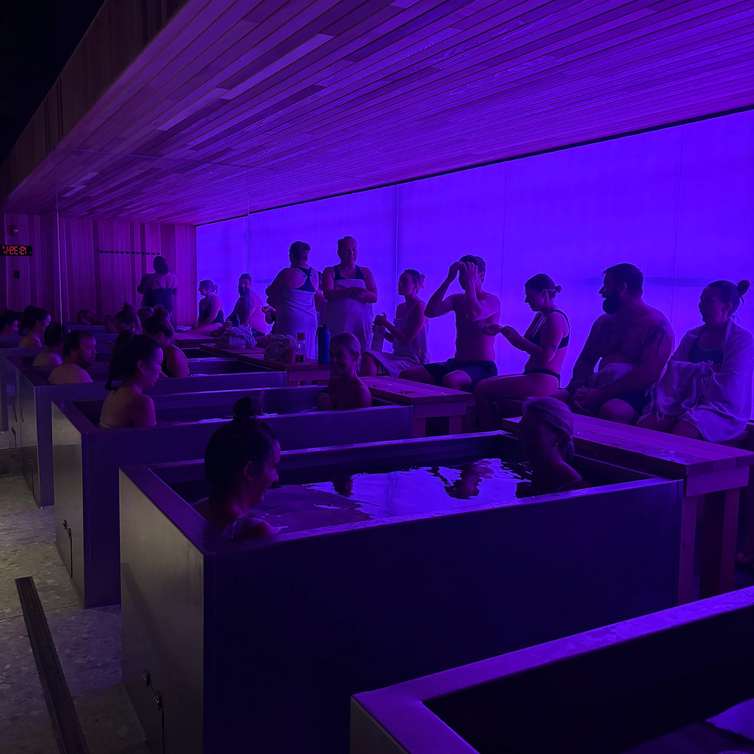 People in a dark, bluish-lit spa with individual hot tubs, some sitting in the tubs and others sitting on chairs or standing, engaging in conversations.