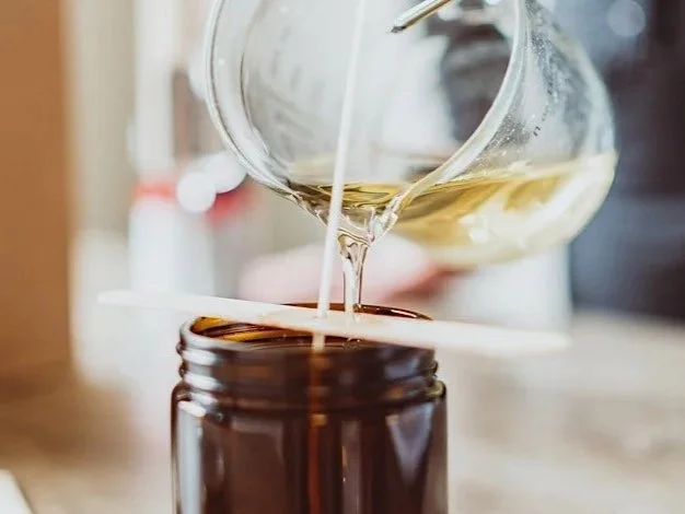 A glass container pouring a clear liquid into a dark brown bottle on a wooden surface.
