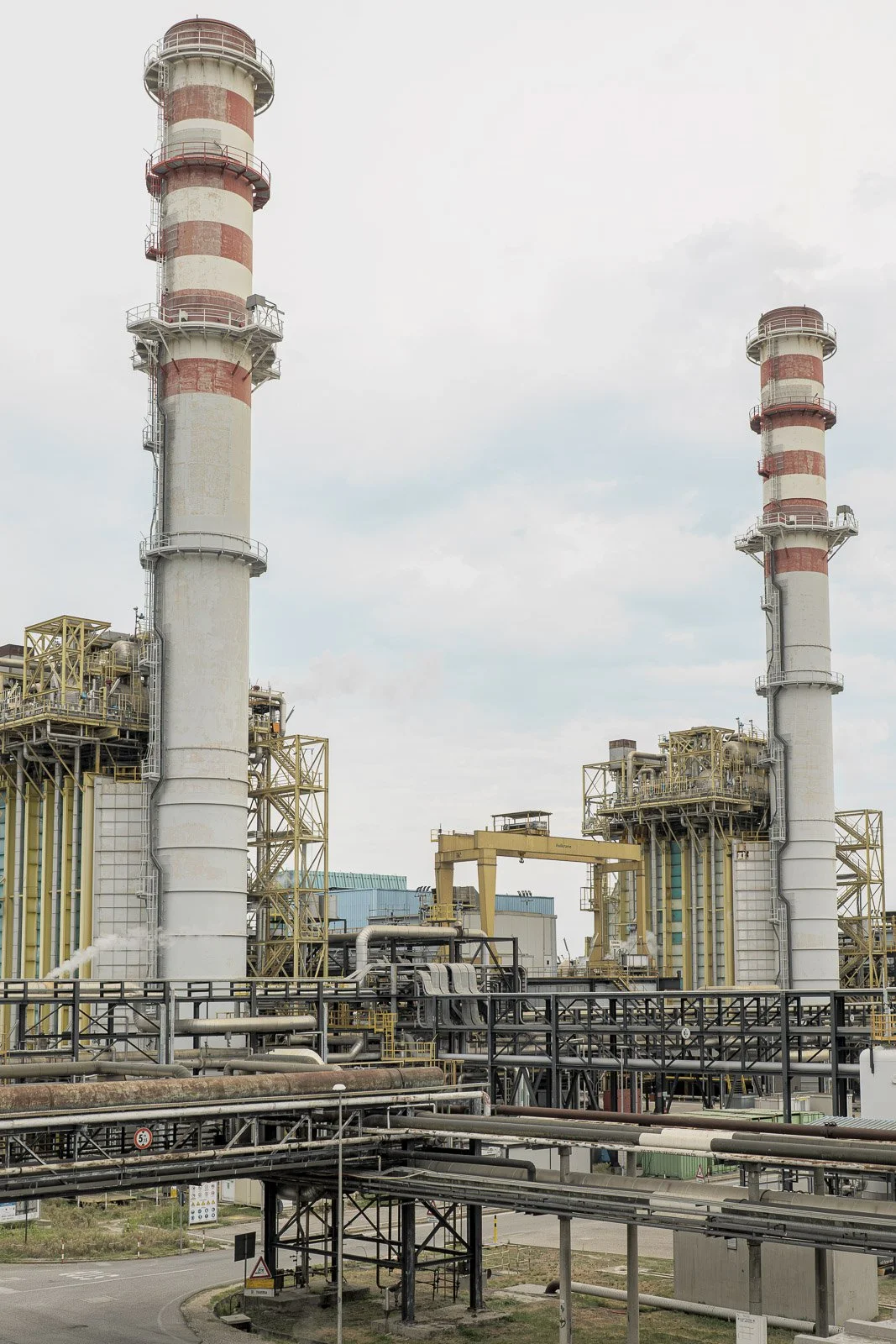 Due ciminiere a fasce bianche e rosse, con tubazioni e strutture metalliche dello stabilimento in primo piano.