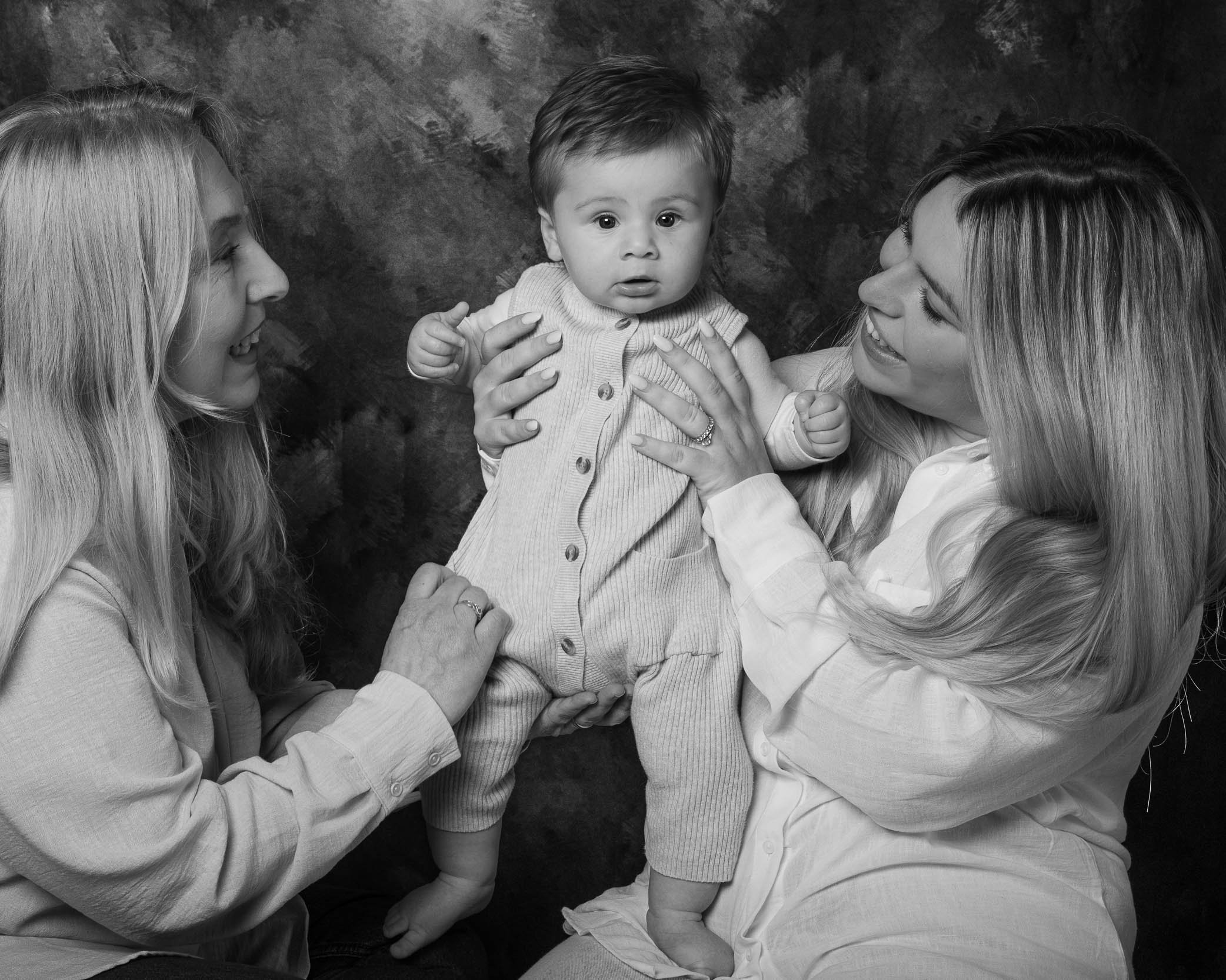 Ritratto di famiglia in studio in bianco e nero con due donne che tengono in braccio un bambino su sfondo artistico – fotografo ritratti a Buccinasco, Milano Sud.
