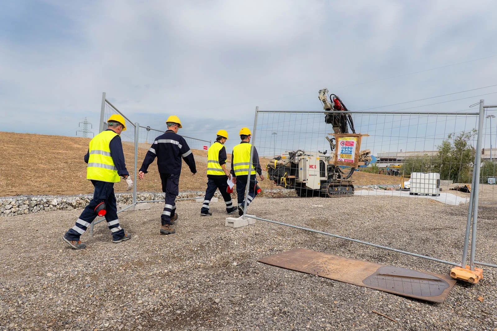 Quattro operatori attraversano l’ingresso recintato del cantiere, dirigendosi verso una perforatrice.