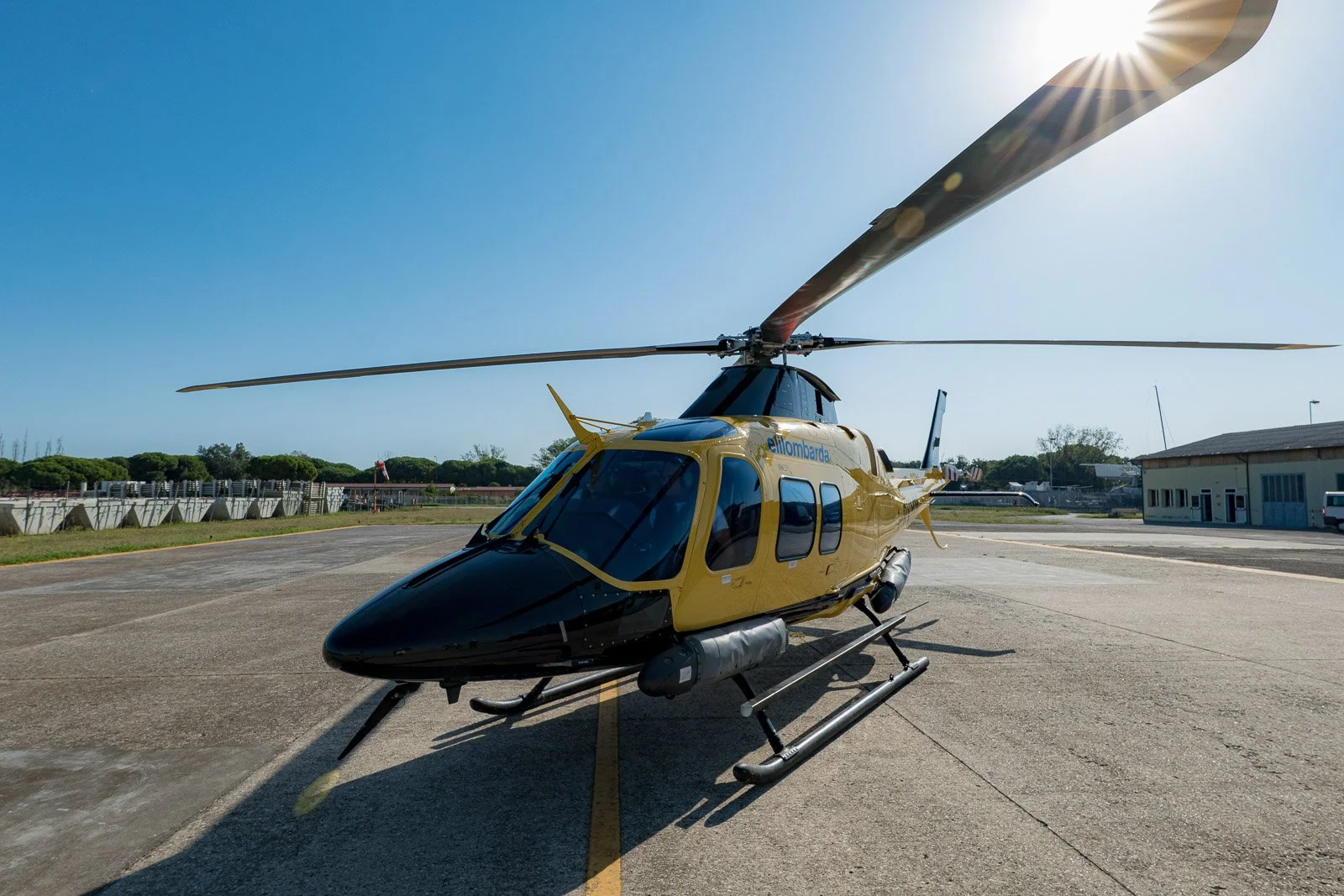 Elicottero giallo e nero Elilombarda sul piazzale, con il sole che filtra dietro una pala del rotore.