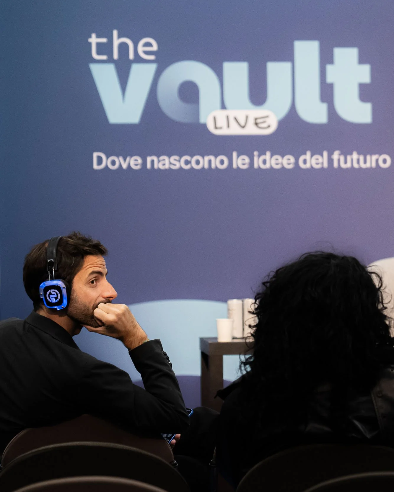 Partecipante con cuffie wireless segue il talk di The Vault Live al MiCo di Milano, davanti al backdrop ufficiale dell’evento organizzato da Will Media e Chora Media.
