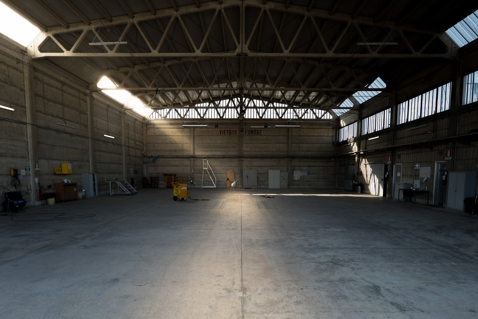 Interno dell’hangar con capriate a vista e fasci di luce naturale che illuminano il pavimento.