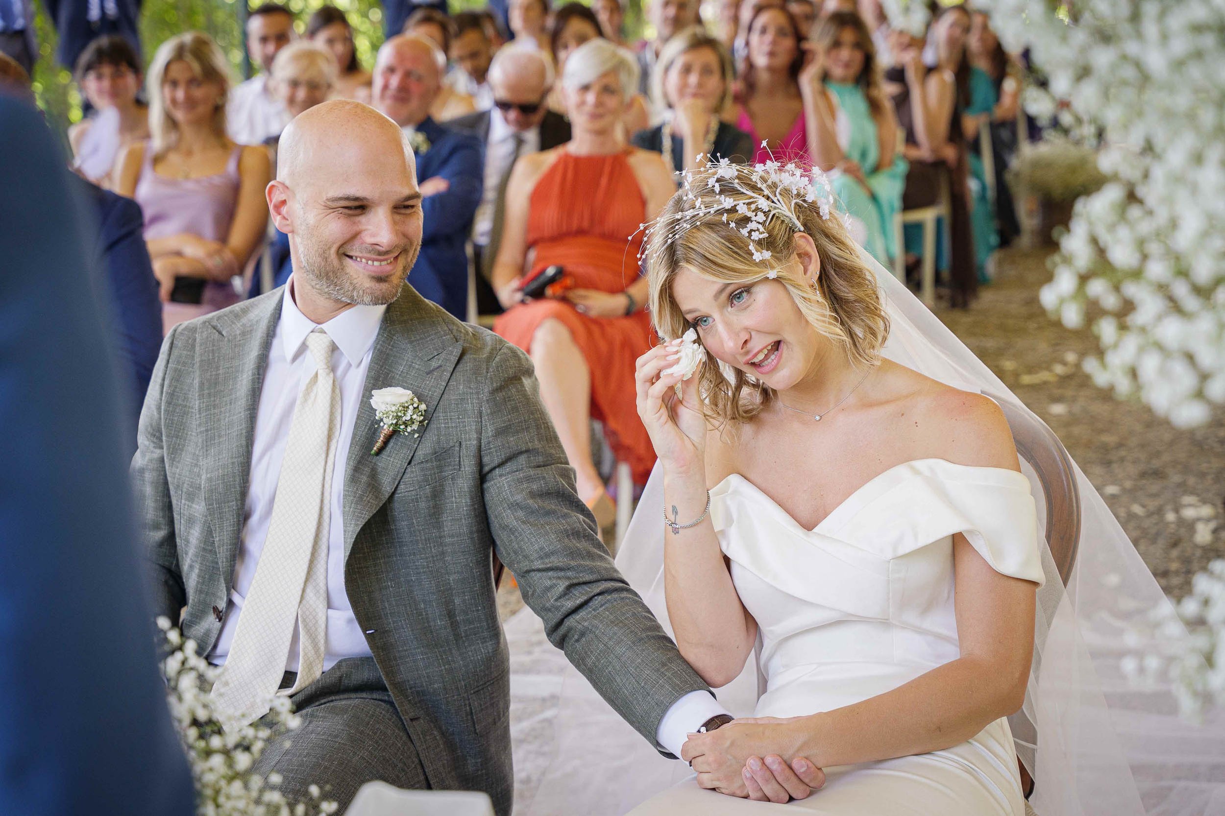 Sposa emozionata durante la cerimonia all’aperto a Tenuta Montezeglio Invorio, mentre asciuga una lacrima tenendo la mano dello sposo