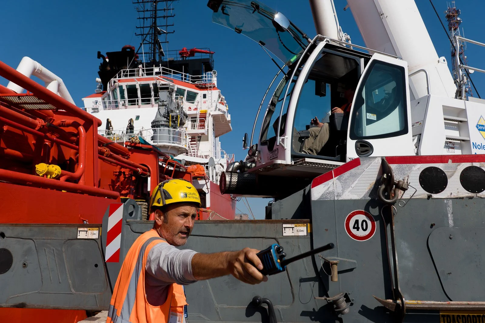Operatore con casco giallo e radio portatile accanto all’autogru, con la nave Asso Ventiquattro sullo sfondo.