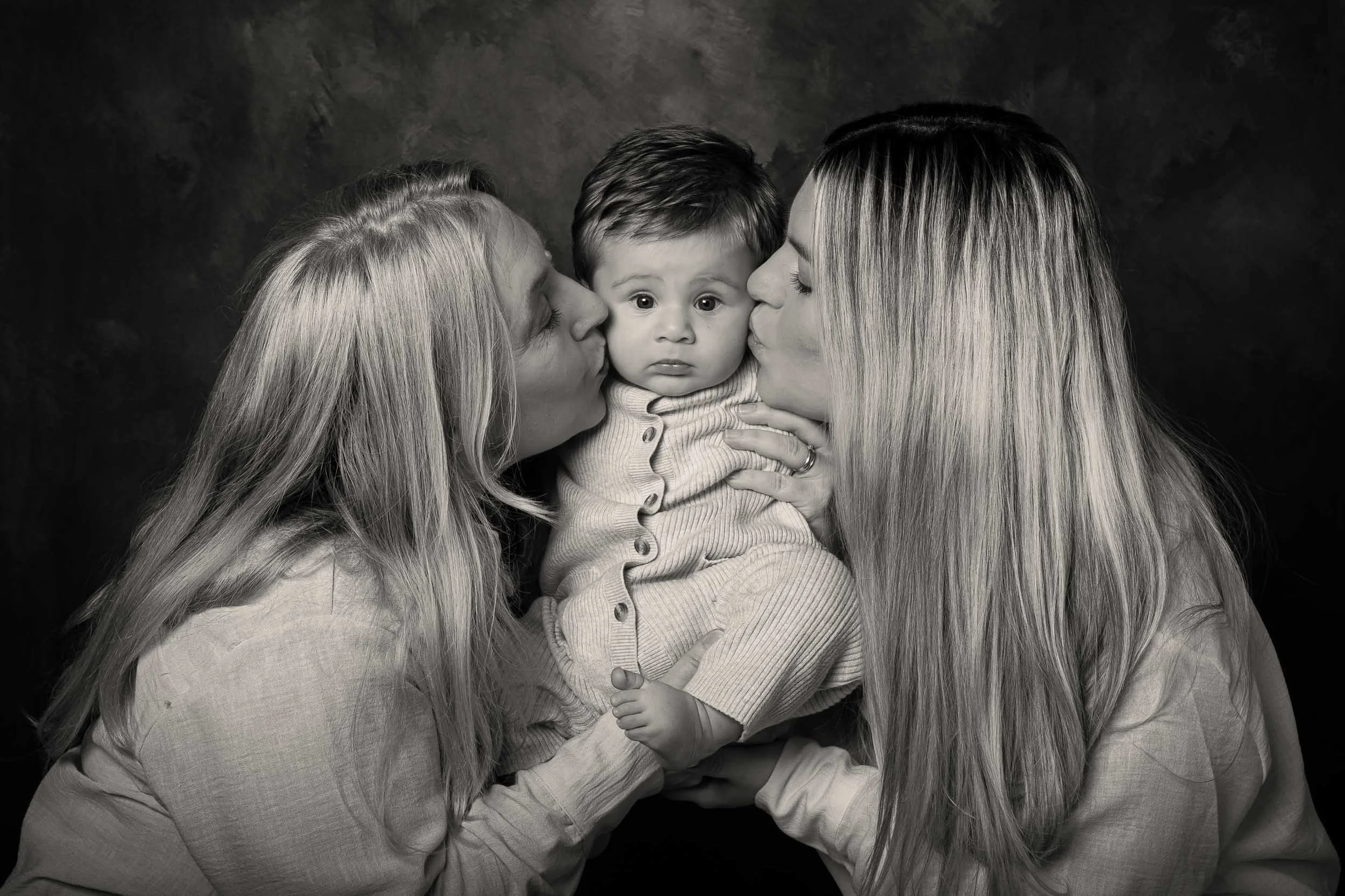 Ritratto di famiglia in studio in bianco e nero con due donne che baciano un bambino al centro su sfondo artistico – servizio fotografico professionale a Buccinasco, Milano Sud.