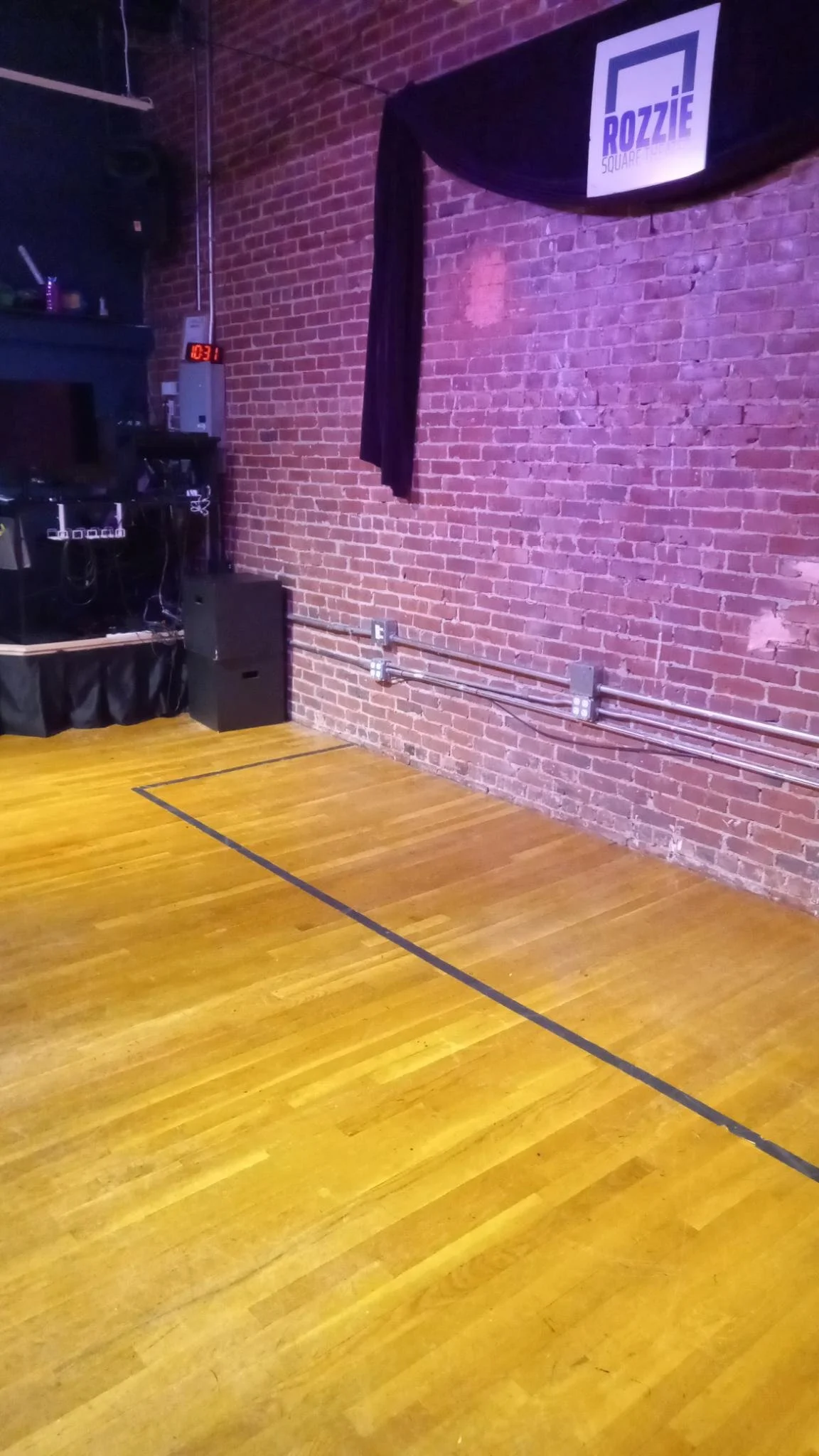 inside a room with a wooden floor marked with tape on the ground, and a brick wall with a curtain hung against it, and some black wooden boxes stacked to the side, next to a wooden platform with computer equipment on it
