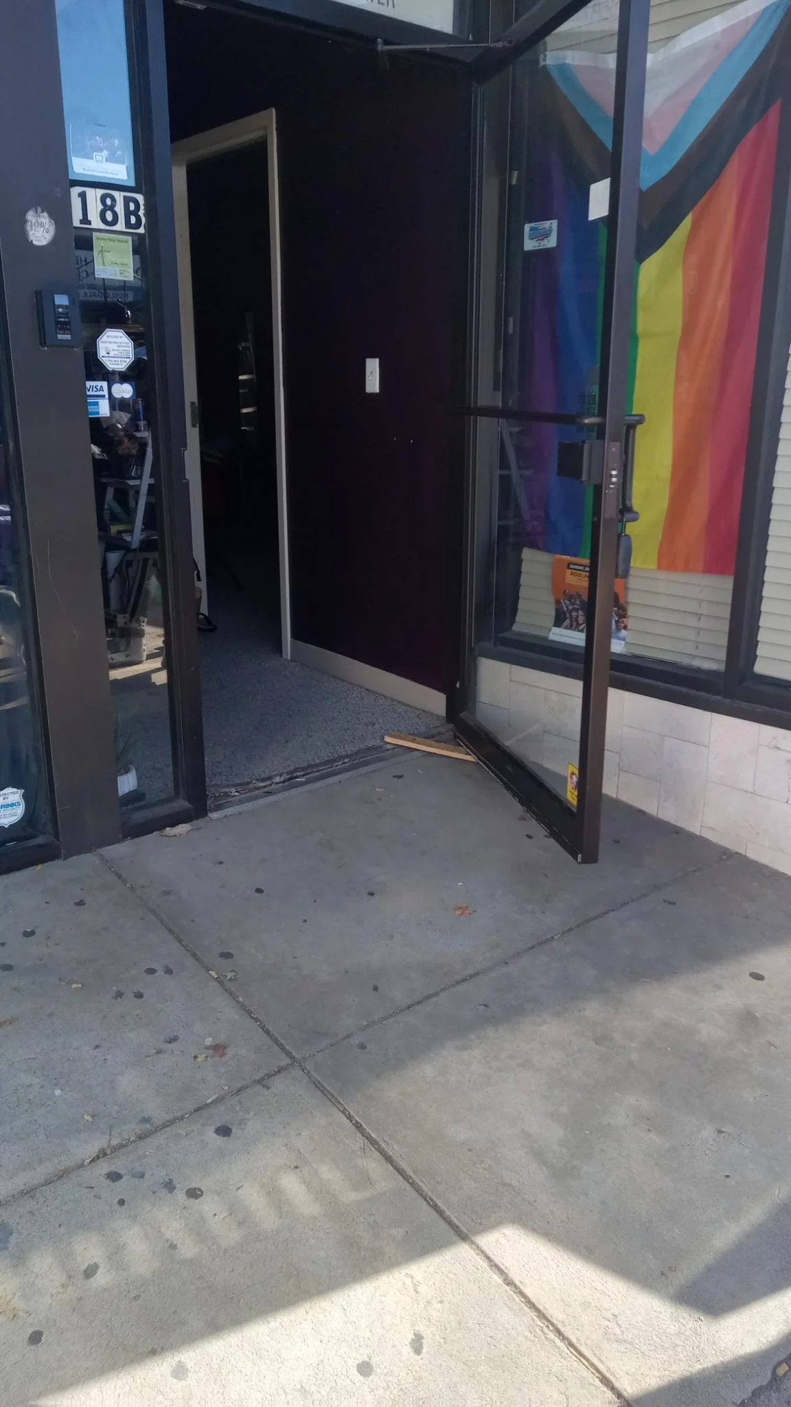 a glass door propped open in front of a rainbow flag, led up to by a sloped pavement ramp