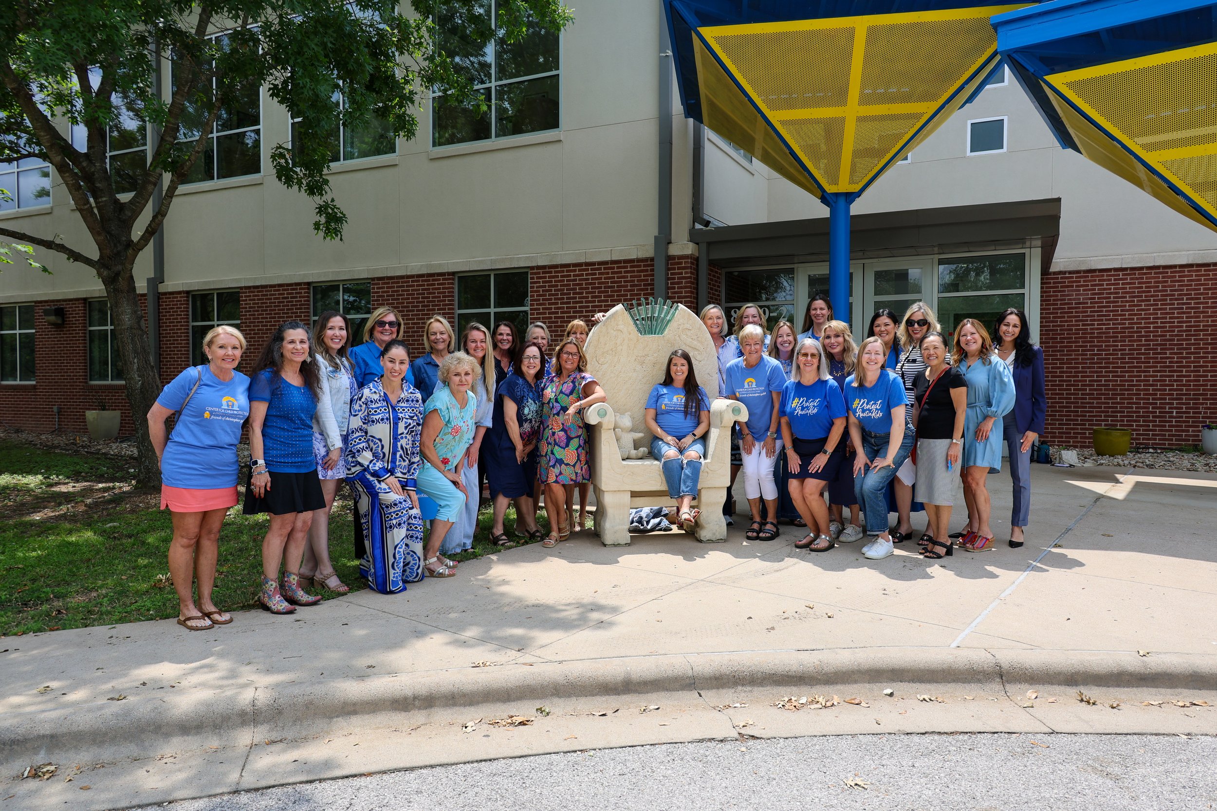 A group of people posing outside a building, with a woman sitting on a large stone chair. Most are wearing blue-themed clothing.