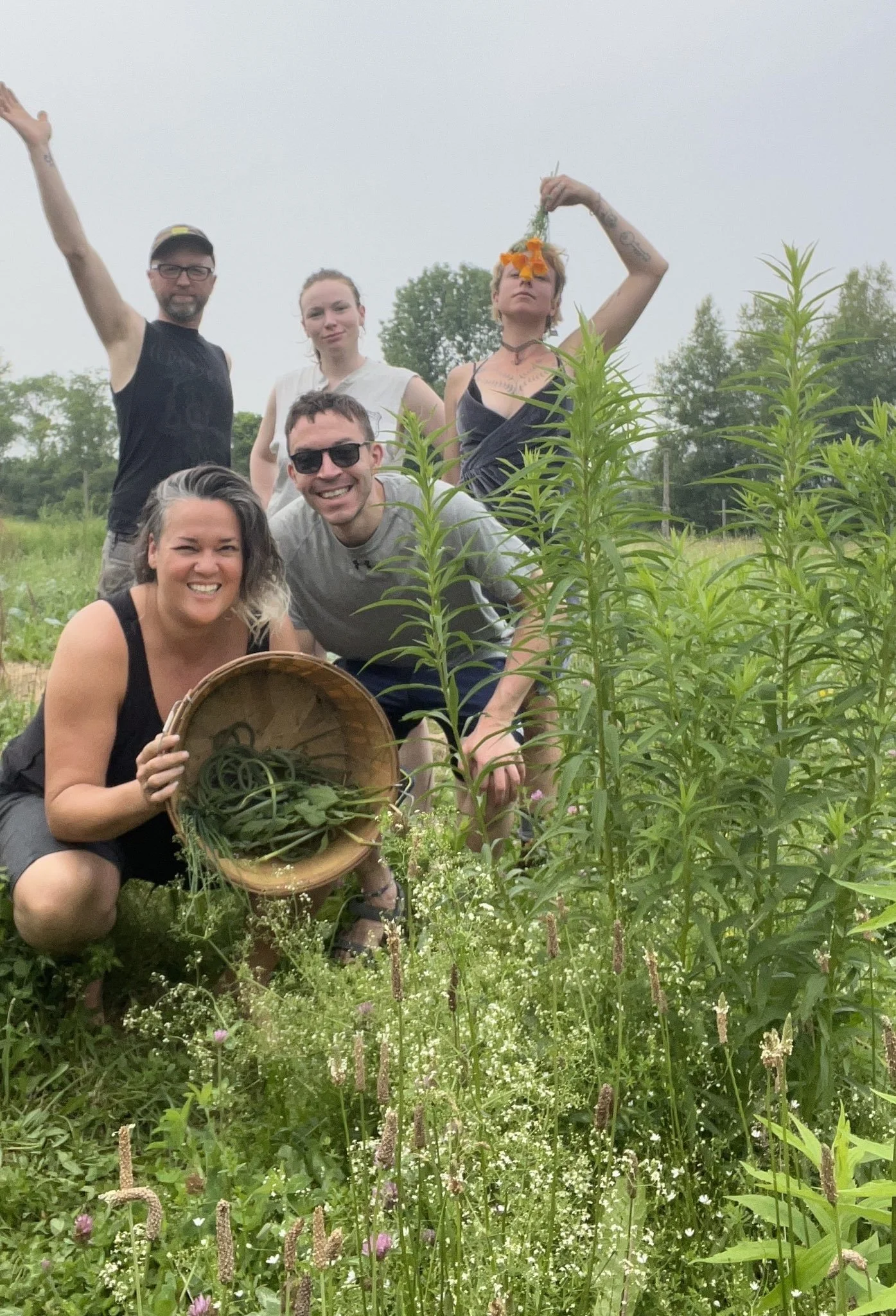 a small group of five smiling people stand in a variety of poses, two squatting holding a basket full of greens, one with an arm out in victory, one with sassy hands on hips, and one with an orange flower to block their face stand in green plants