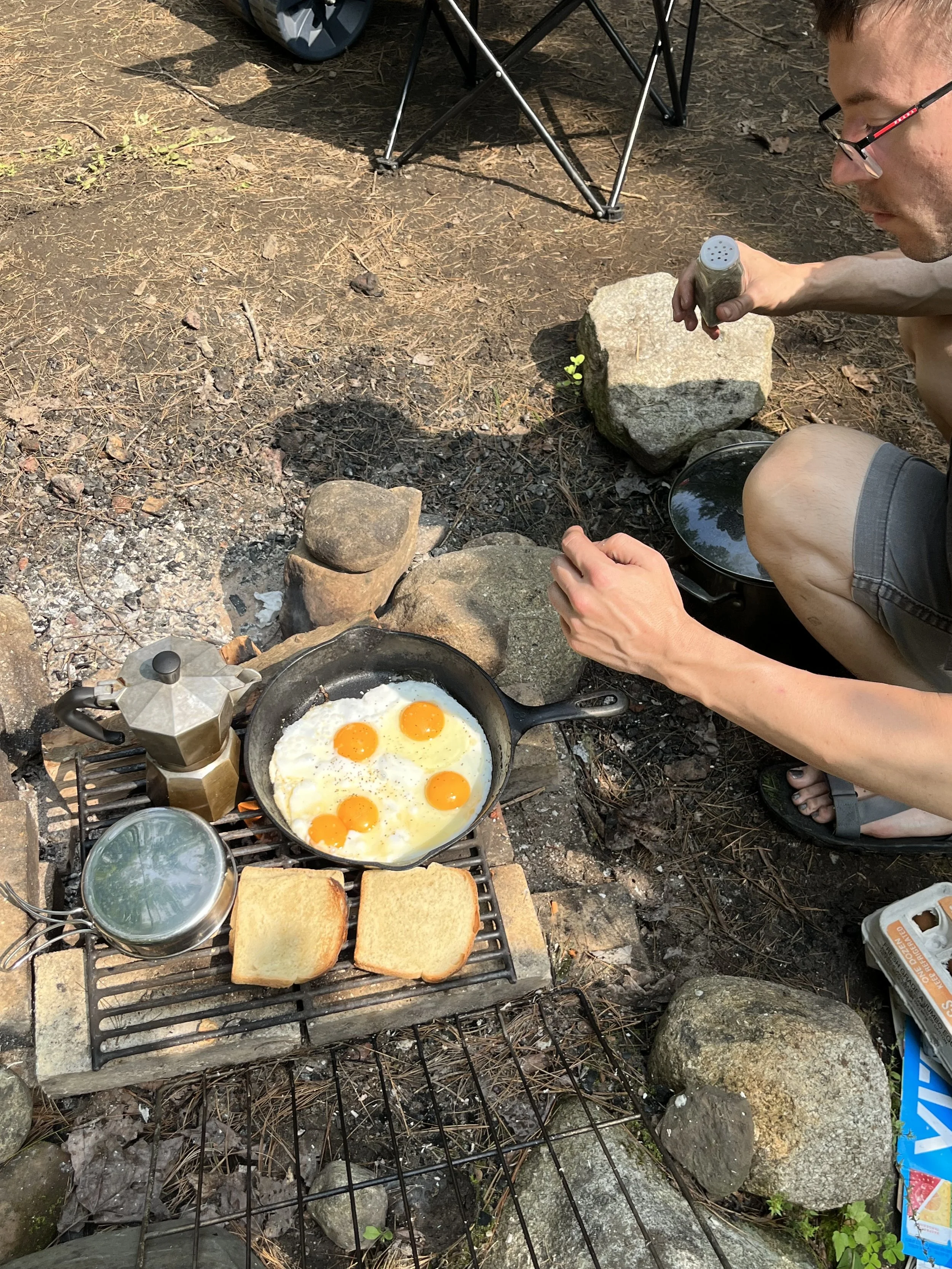 A person squats next to a fire pit with a pan full of 5 sunny side up eggs. There is toast slices on the grill and a pot of coffee all heating up. There is a camping chair leg in the corner