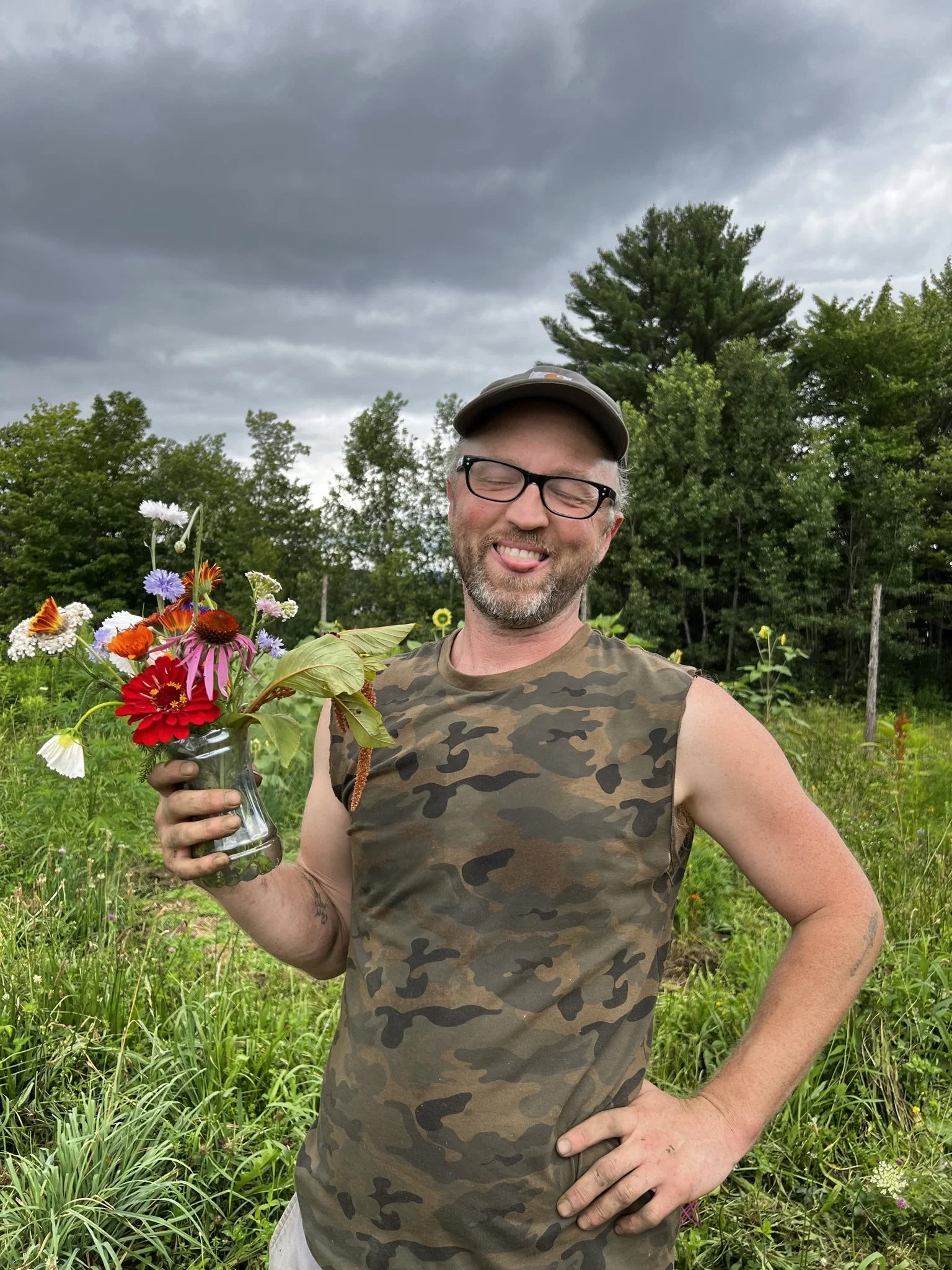 a goofy smiling man in a hat and camouflage t-shirt holds a bouquet of garden flowers in front of a field of greens
