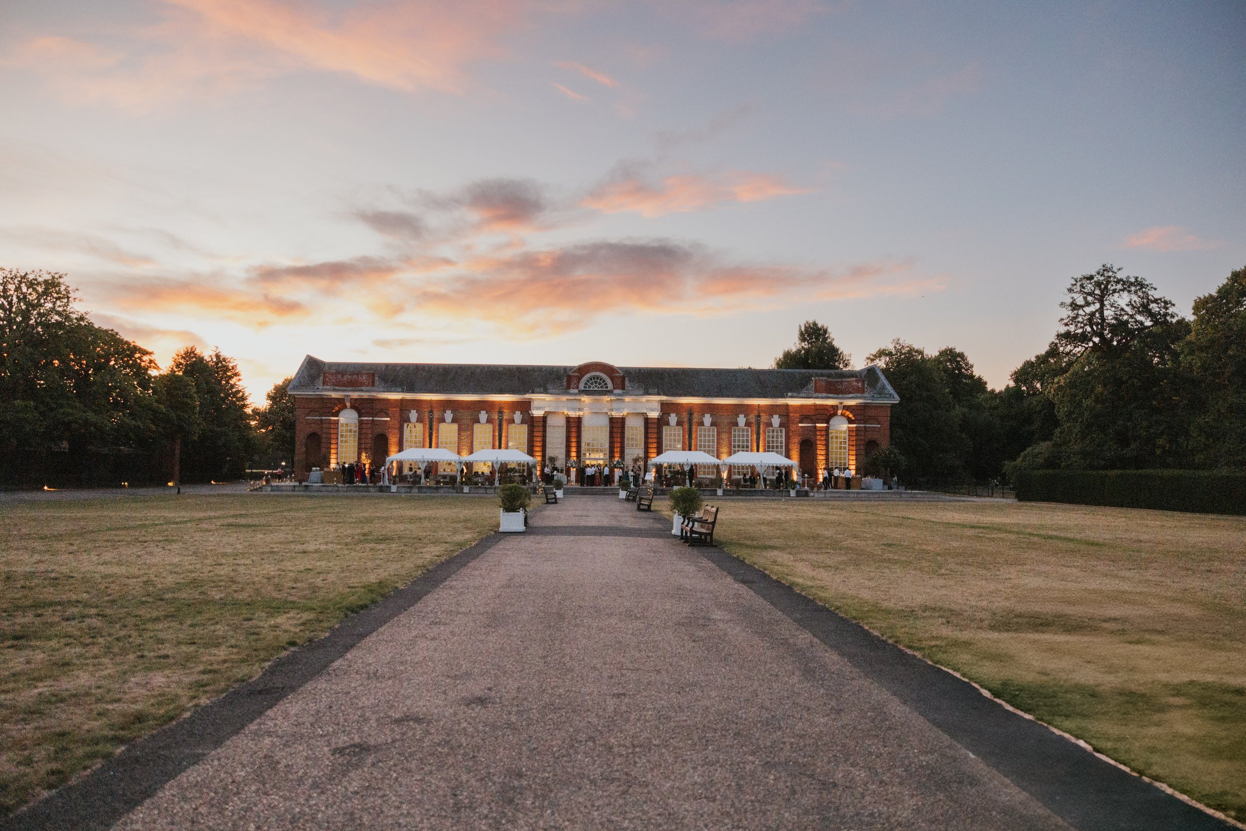 Drinks reception at dusk on the grounds of Kensington Palace with guests mingling in the formal gardens and the palace visible behind them