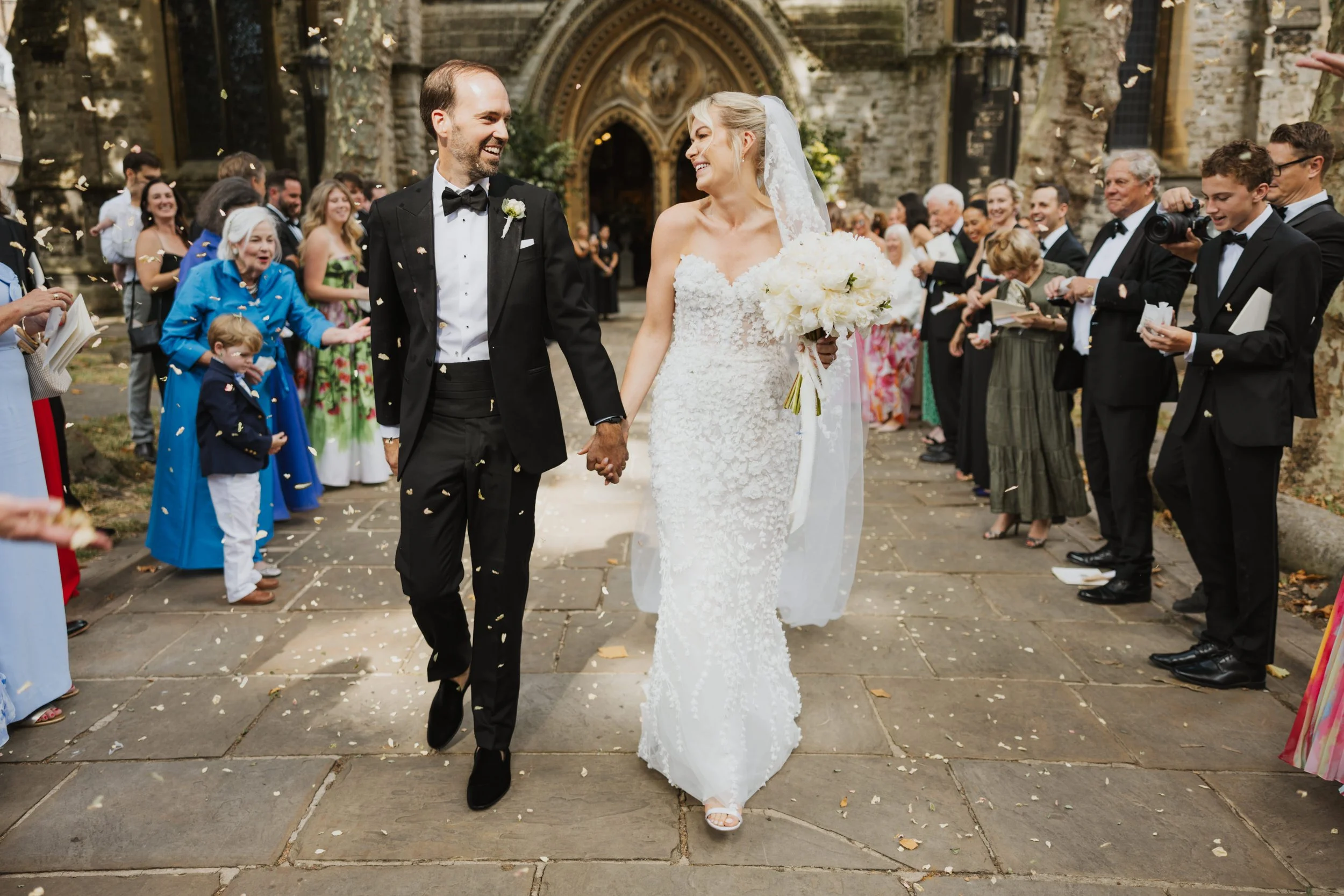 Mimi and Simon exiting their St Abbots Kensington church ceremony through a shower of confetti with guests cheering outside